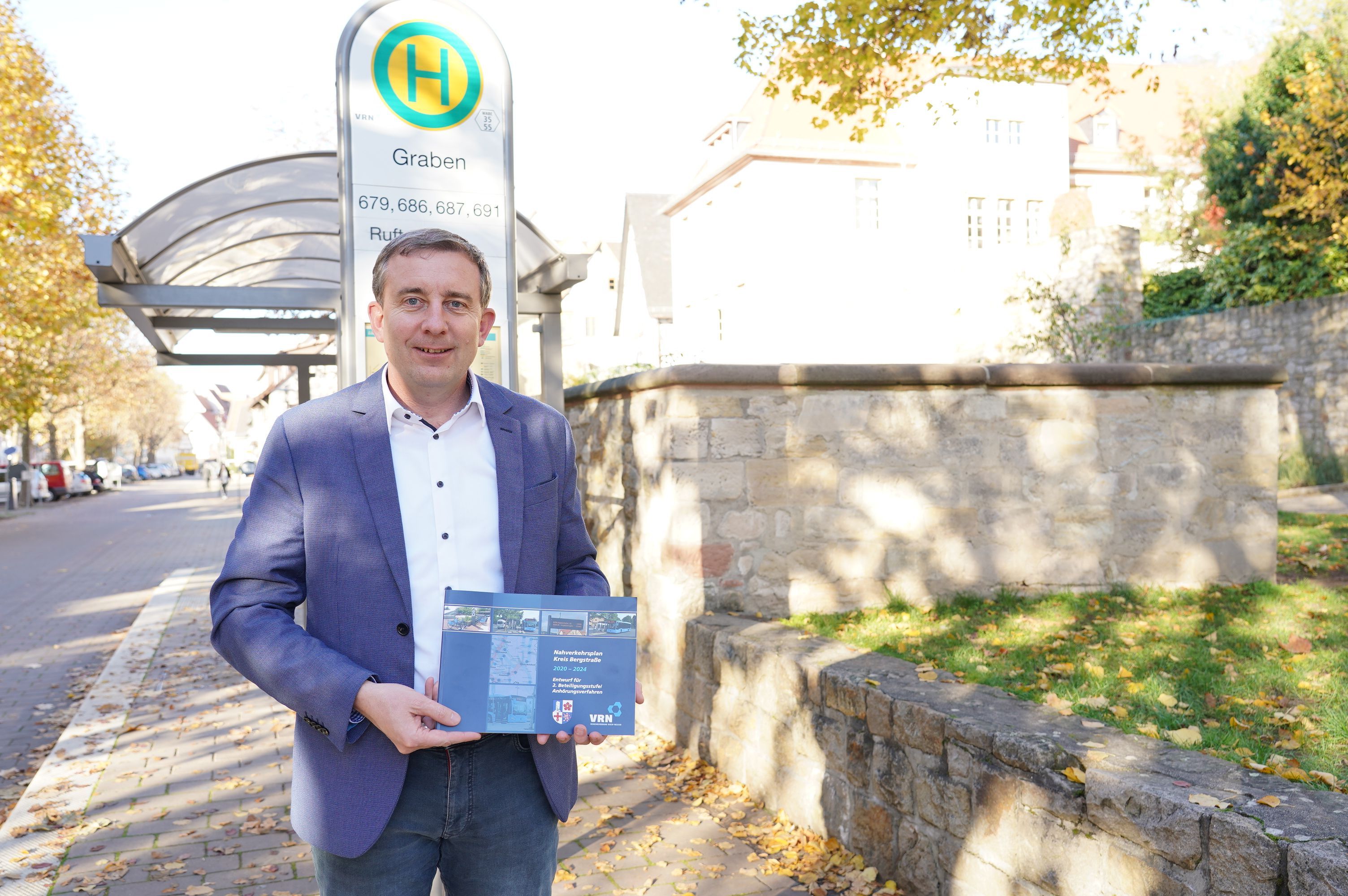 Karsten Krug steht vor einer Bushaltestelle und hält den Nahverkehrsplan in den Händen. 