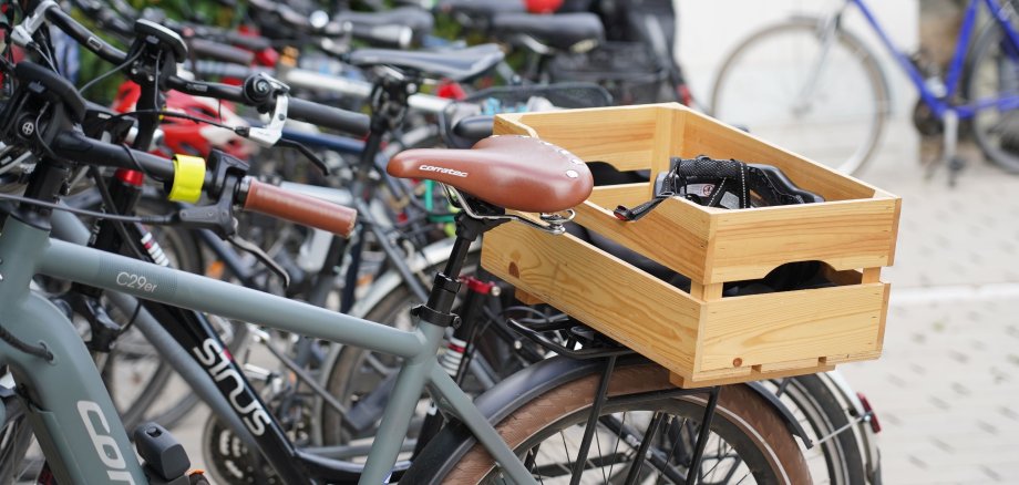 Blick auf eine Reihe von Fahrrädern, die nebeneinander stehen. Das vorderste Rad hat eine Holzkiste als Korbersatz auf dem Gepäckträger