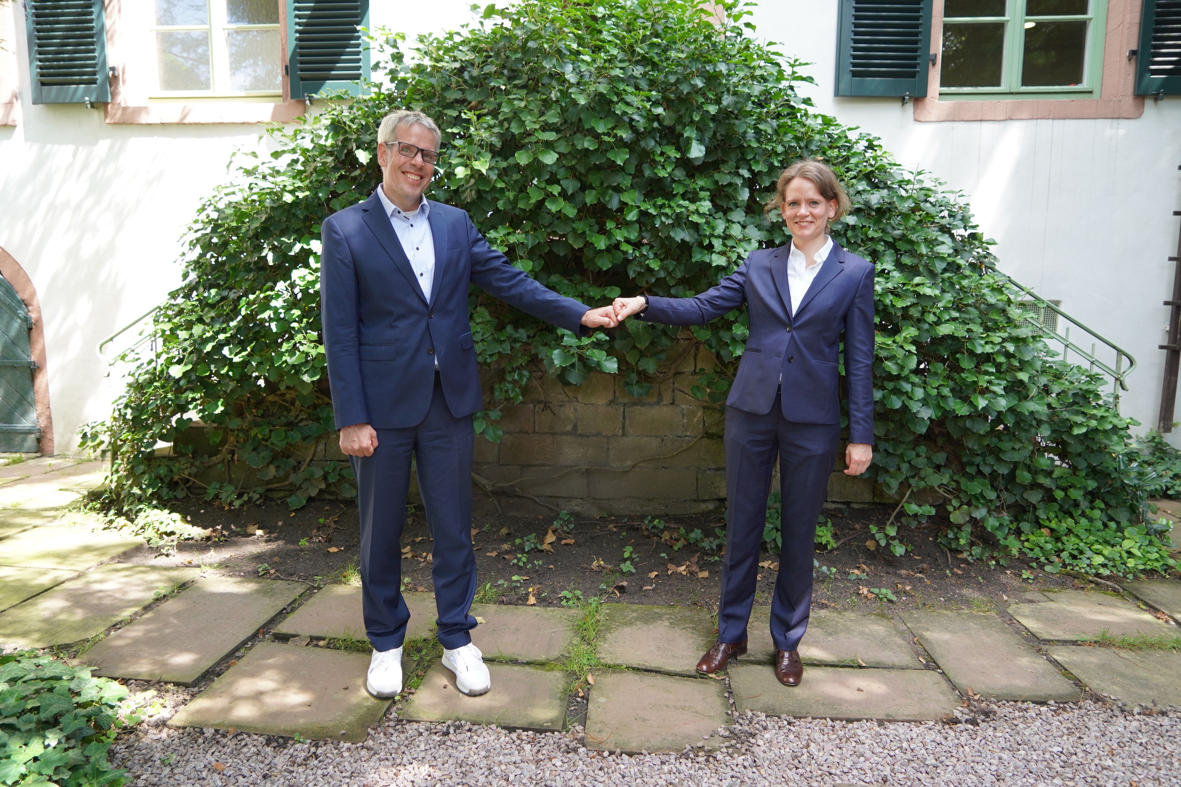Landrat Christian Engelhardt und die Polizeidirektorin Juliane Ries stehen vor einer mit Efeu bewachsenen Treppe und halten die Fäuste aneinander