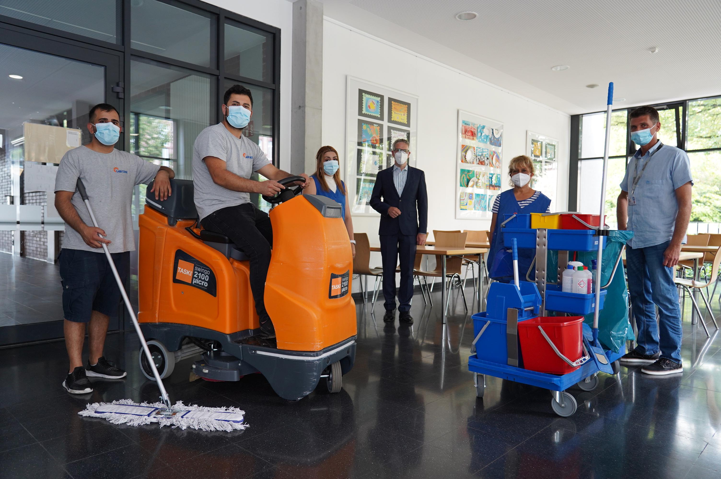 Landrat Christian Engelhardt steht in der Mitte von fünf Personen. Der mann ganz rechts hält einen Wischmopp in der Hand, der Mann daneben sitzt auf einem orangenen Gefährt. Ganz recht außen steht ein Vertreter von LSG in Alltagsklamotten, neben ihm eine Frau mit einer blauen Schürze. Vor den beiden steht ein Putzwagen.