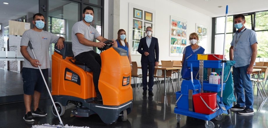 Landrat Christian Engelhardt steht in der Mitte von fünf Personen. Der mann ganz rechts hält einen Wischmopp in der Hand, der Mann daneben sitzt auf einem orangenen Gefährt. Ganz recht außen steht ein Vertreter von LSG in Alltagsklamotten, neben ihm eine Frau mit einer blauen Schürze. Vor den beiden steht ein Putzwagen.