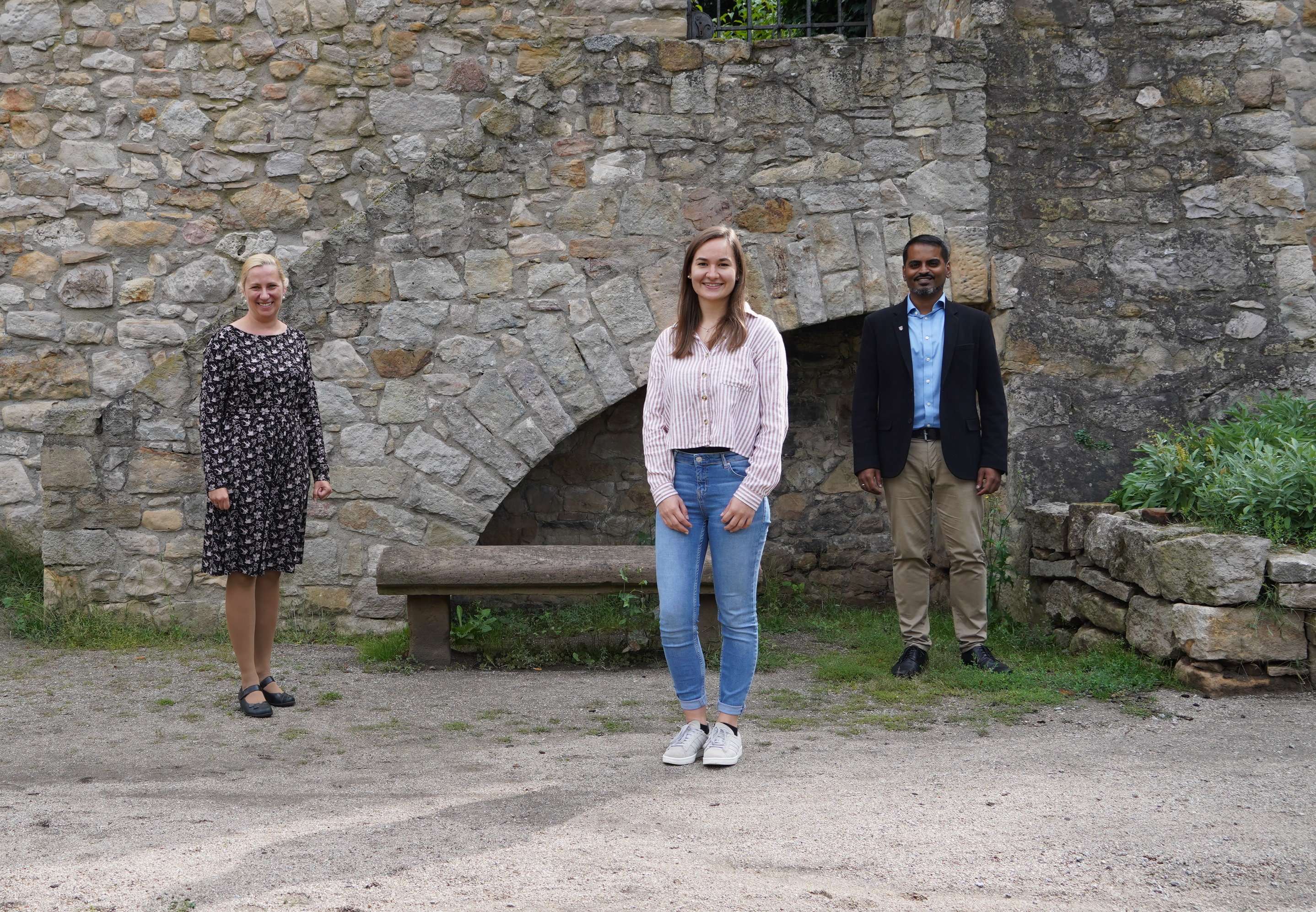 Vor einer alten Steinmauer steht im Vordergrund eine sportlich gekleidete junge Frau, die Hebamme Marie Schmitt. Links daneben, einen Schritt nach hinten Versetzt, steht die Gesundheitsdezernentin Diana Stolz. Auf der rechten Seite, auf Höhe von Daana Stolz, steht der Birkenauer Bürgermeister Milan Mapplassary
