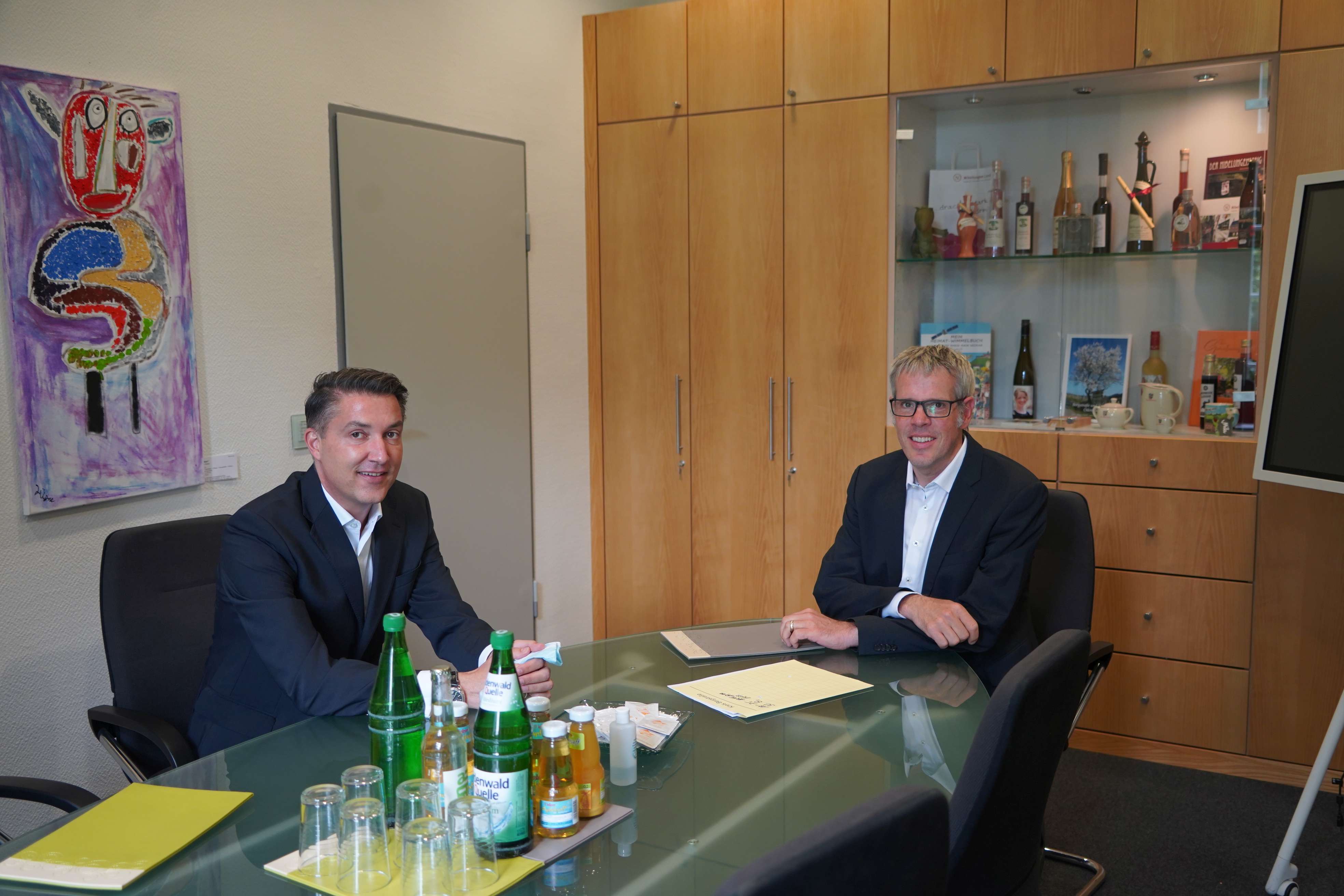 An einem großen Glastisch, auf dem Akten und eine Auswahl an Getränken zu sehen sind, sitzen der Landrat und Johannes Erich Schulz und lächeln in die Kamera.