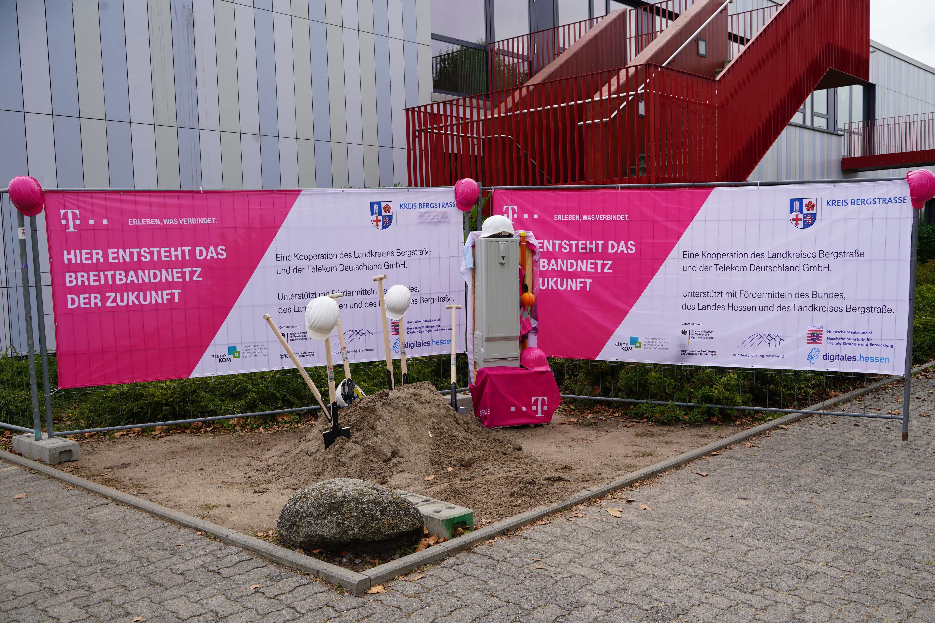 Blick auf zwei Bauzäune, an welchen magentafarbene Telekom-Banner mit dem Wappen des Kreis Bergstraße hängen. Vor den Bauzäunen ist ein kleiner Erdhügel mit Schaufeln und Helmen zu sehen.