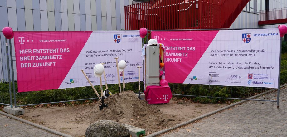 Blick auf zwei Bauzäune, an welchen magentafarbene Telekom-Banner mit dem Wappen des Kreis Bergstraße hängen. Vor den Bauzäunen ist ein kleiner Erdhügel mit Schaufeln und Helmen zu sehen.