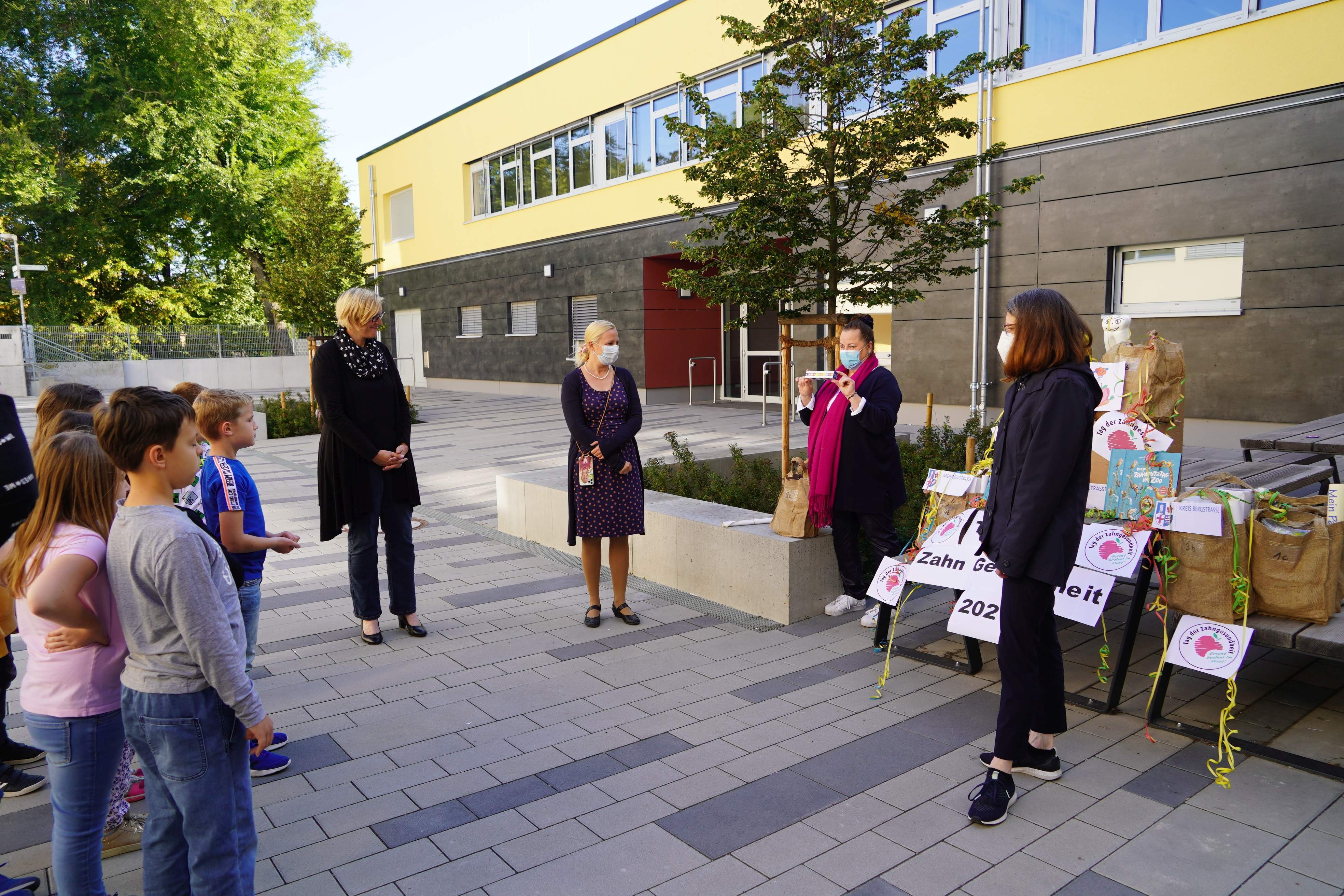 Zu sehen sind vier Frauen, die einer Gruppe Grundschülern am linken Bildrand etwas über das Thema Zahnhygiene erklären.