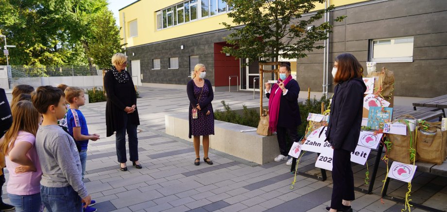 Zu sehen sind vier Frauen, die einer Gruppe Grundschülern am linken Bildrand etwas über das Thema Zahnhygiene erklären.