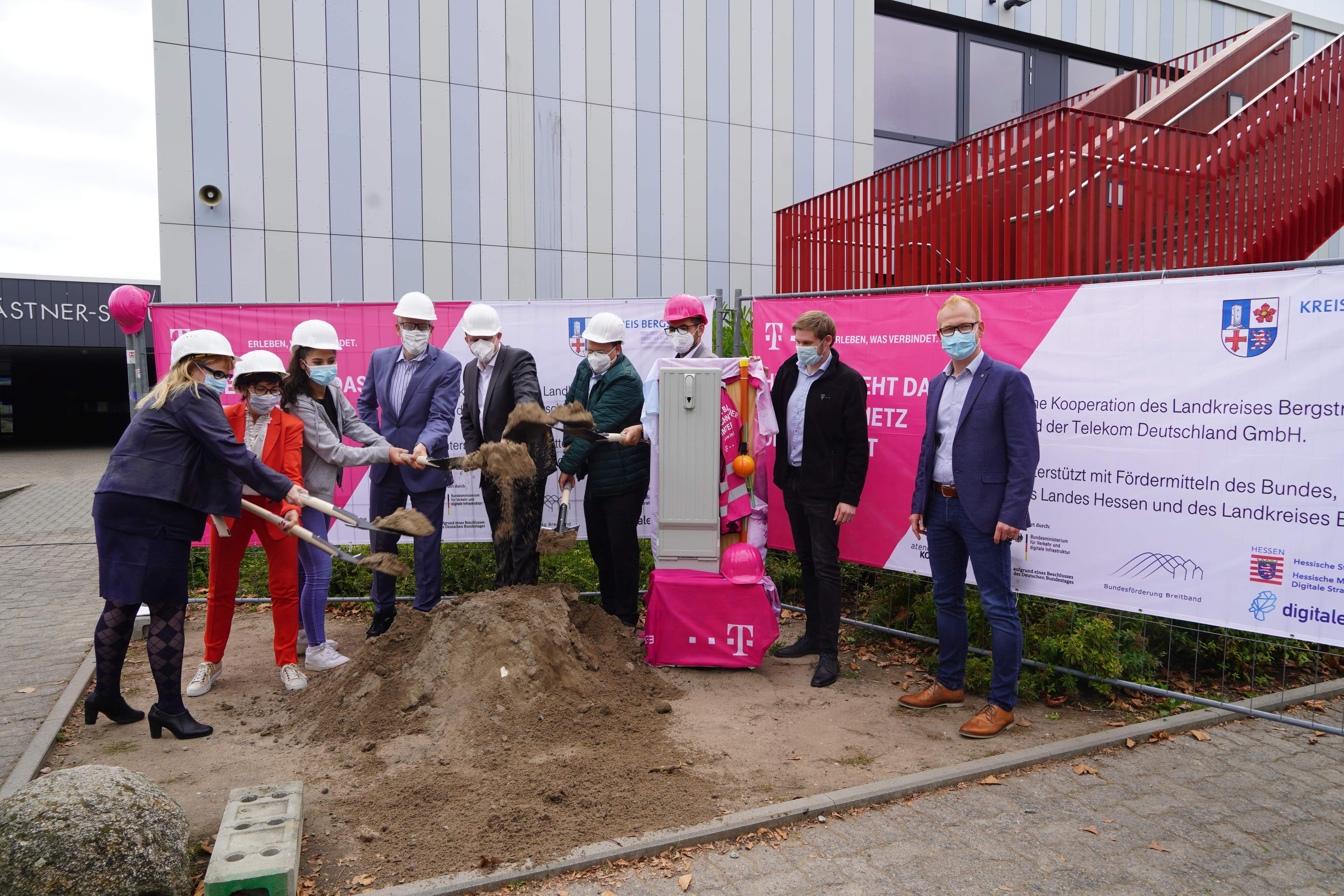 Ein Gruppenbild vor magentafarbenen Bannern der Telekom bei dem ein Großteil der Personen Erde mein Spaten in die Luft wirft und weiße Helme trägt. 