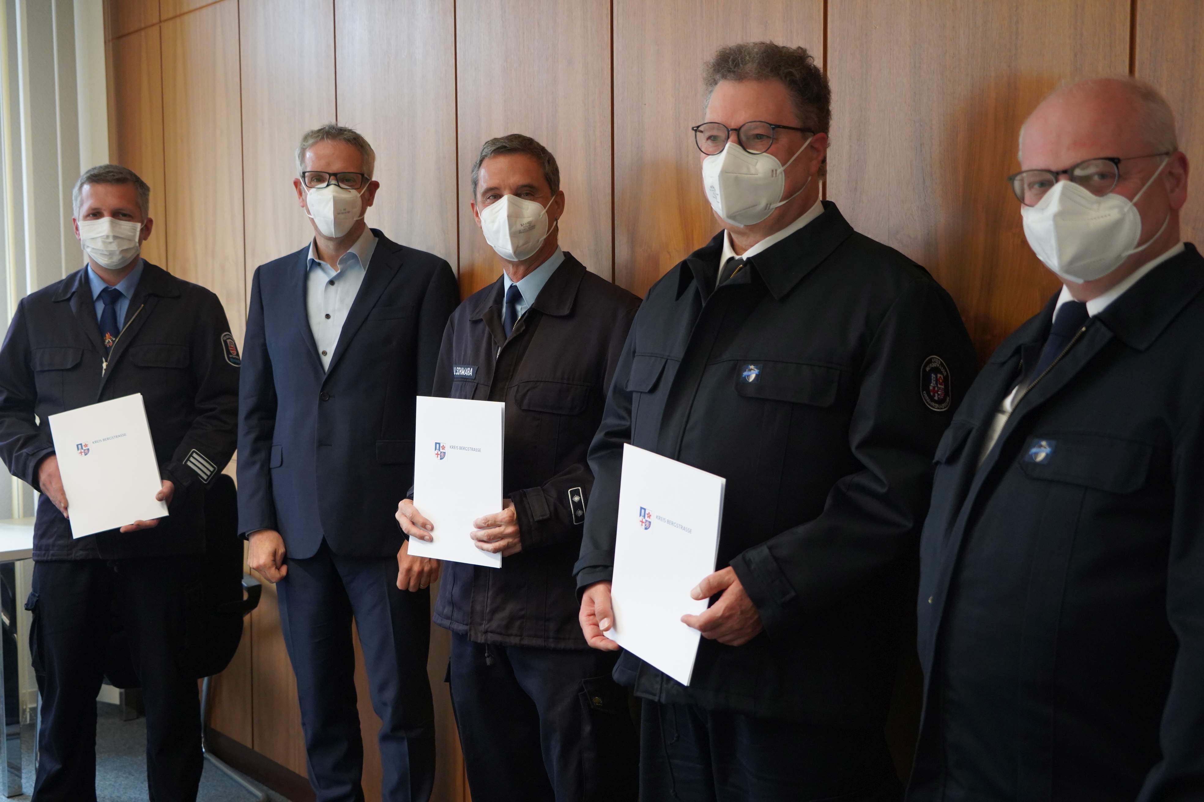 Landrat Christian Engelhardt und Kreisbrandinspektor Steffen Lutter bei der Vereidigungszeremonie für die neuen Kreisbrandmeister Bastian Weisener, Uwe Schwara und Karl-Christian Hahn. All vier fünd Männer sind dunkel gekleidet, drei von ihnen halten weiße Mappen in den Händen.