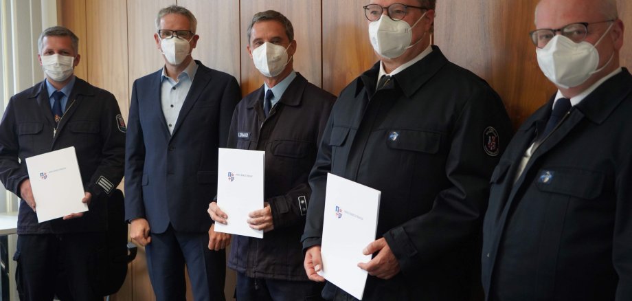 Landrat Christian Engelhardt und Kreisbrandinspektor Steffen Lutter bei der Vereidigungszeremonie für die neuen Kreisbrandmeister Bastian Weisener, Uwe Schwara und Karl-Christian Hahn. All vier fünd Männer sind dunkel gekleidet, drei von ihnen halten weiße Mappen in den Händen.