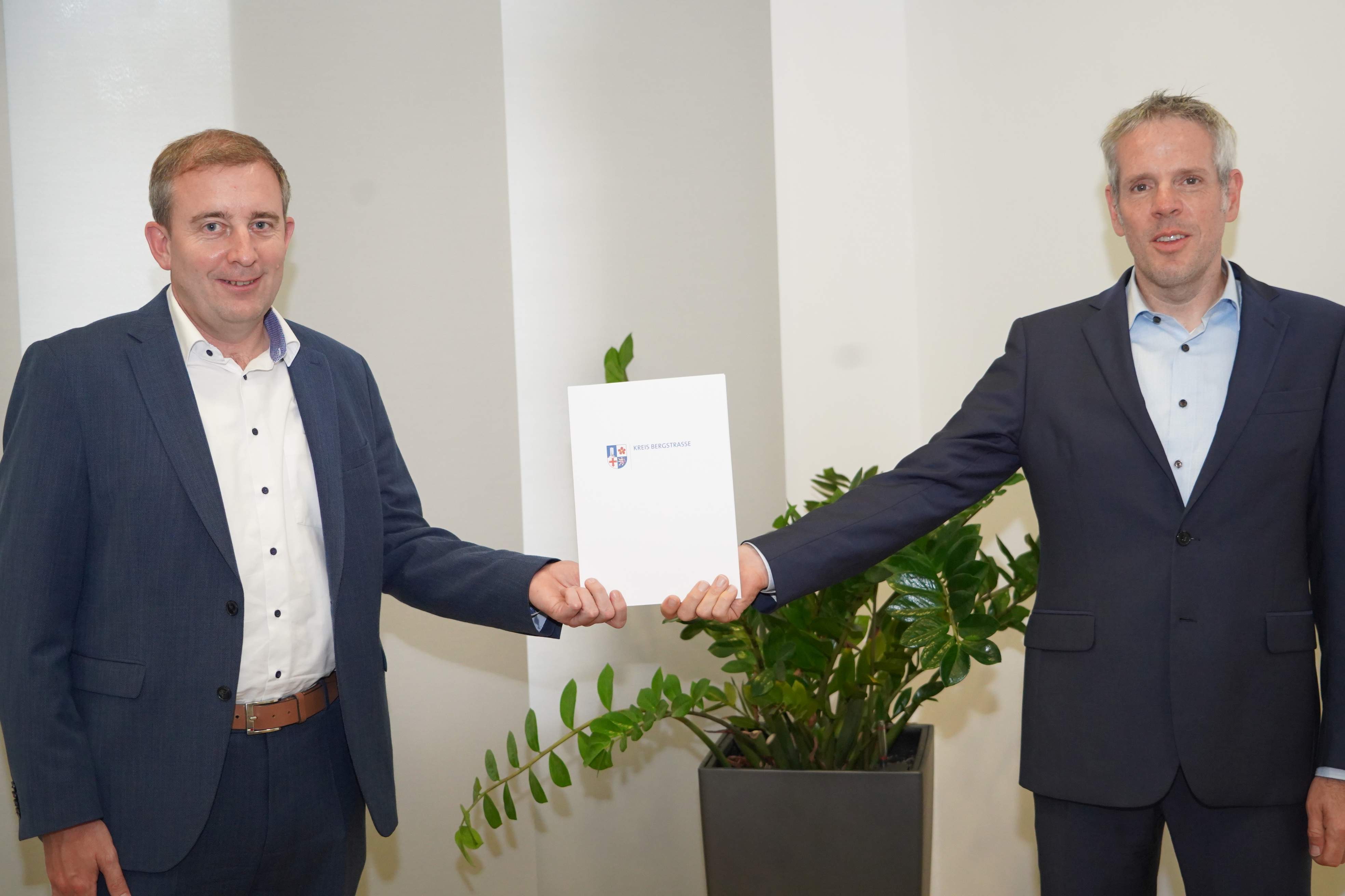 Landrat Christian Engelhardt und Karten Krug, beide in dunklen Anzügen, stehen nebeneinander und halten zwischen sich eine weiße Kreiß-Bergstraße-Mapp hoch. 