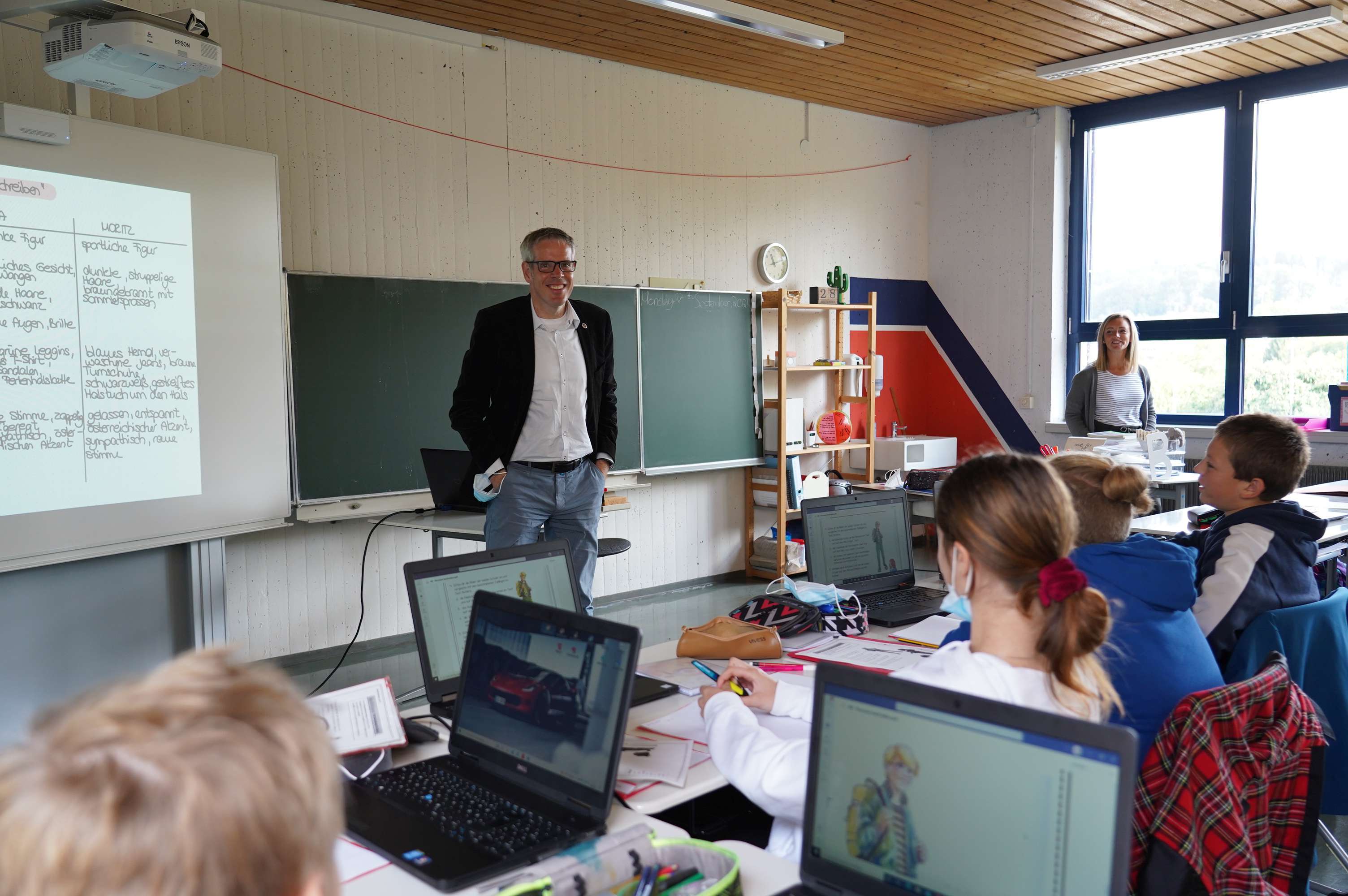 Landrat Christian Engelhard steht in einem Klassenzimmer und lächelt in die Kamera. Auf den Tischen der Schülerinnen und Schüler stehen Laptops.