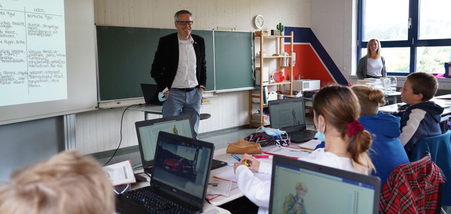 Landrat Christian Engelhard steht in einem Klassenzimmer und lächelt in die Kamera. Auf den Tischen der Schülerinnen und Schüler stehen Laptops.
