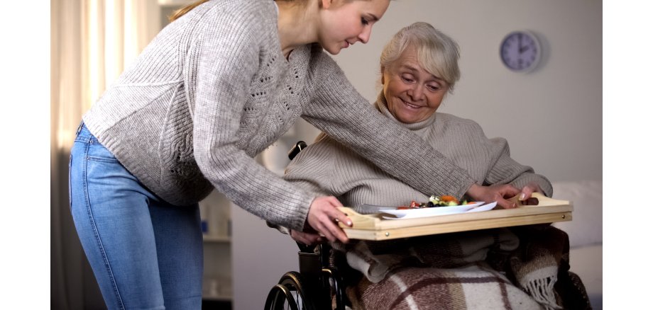 Helferin bringt Seniorin im Rollstuhl ein Essenstablett