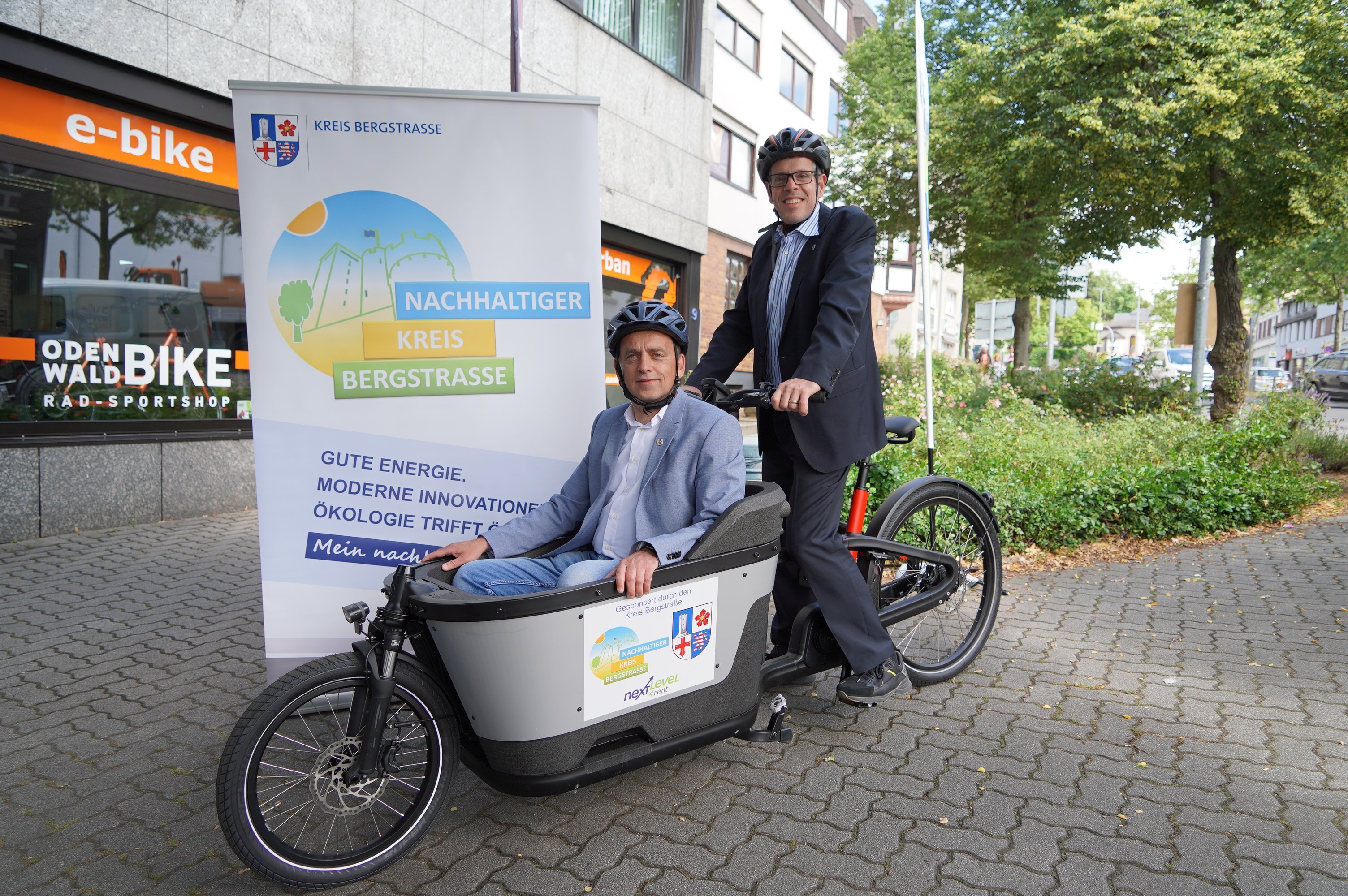 Landrat Christian Engelhardt sitzt auf einem Lastenfahrrad. Im Korb des Rades davor sitzt sein Fahrer, Gerd Oravecz. Beide testen das neue Lastenradangebot.