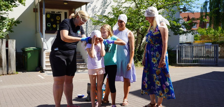 Auf dem Bild zu sehen sind die  Gesundheitsdezernentin Diana Stolz, die Lehrerin Caroline Schmitt und Ute Fuchs vom Bergsträßer Gesundheitsamt. Sie helfen den Schülerinnen Nina Janowska (vorne) und Pia Meiswinkel beim Anlegen eines „Piraten-Sonnenschutzes“ aus Verbandsmaterialien. Auch die drei Erwachsenen tragen den weißen Verband als Sonnenschutz auf dem Kopf.