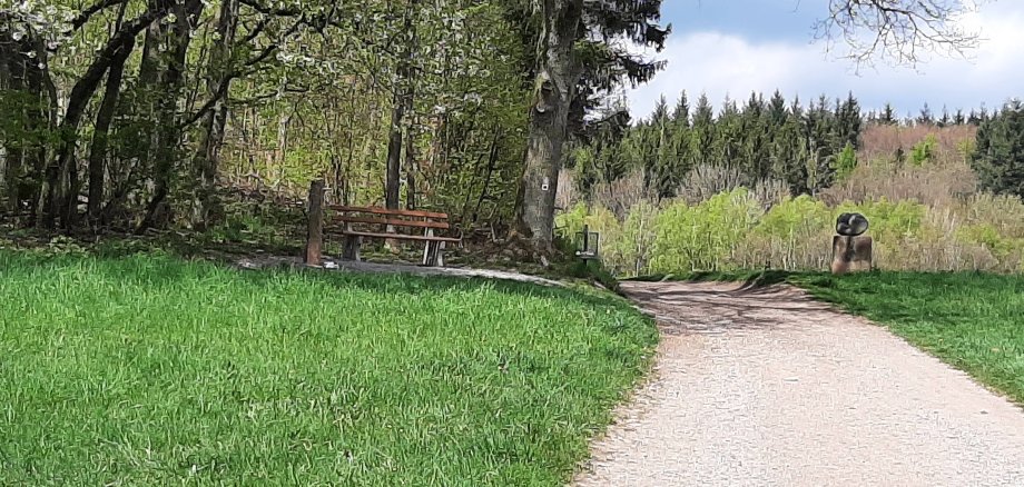 Das Bild zeigt den Kunst-Wanderweg Waldmichelbach. Rund um den Weg sind grüne Wiesen und Wälder, am rechten Wegesrand ist auch eine Stein-Skulptur zu sehen.