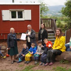 Auf dem Außengelände des Waldkindergartens Wilde Möhre wird ein Förderbescheid überreicht. Vize-Landrätin Diana Stolz (links) überreicht den Bescheid an Thomas Jungfleisch (Geschäftsführer des Vereins KuBus-Kröckelbach, Träger der Kita), mit dabei (von links) Verena Weiß vom Bergsträßer Jugendamt, die Leiterin des Waldkindergartens Nicole Wetzel und Kita-Mitinitiatorin Kathinka Winkler sowie Kindergartenkinder.