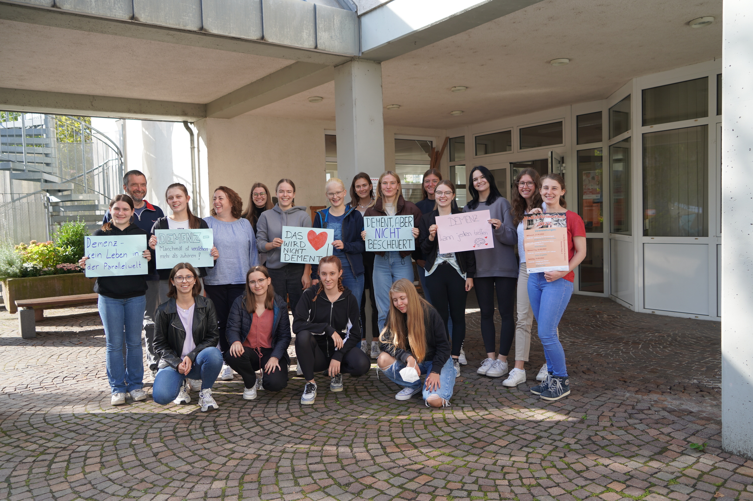 Schülerinnen der Liebfrauenschule in Bensheim stehen gemeinsam mit ihren Tutoren Dr. Nikolas Chalwatzis (2. von links) und Dr. Anette Grüttgen (5. von links) vor ihrer Schule und halten Plakate in die Höhe, die sich mit dem Thema Demenz auseinandersetzen. Darauf stehen Slogans wie etwa „Das Herz wird nicht dement“ oder „Demenz – manchmal sind es kleine Gesten, nicht große Worte“.