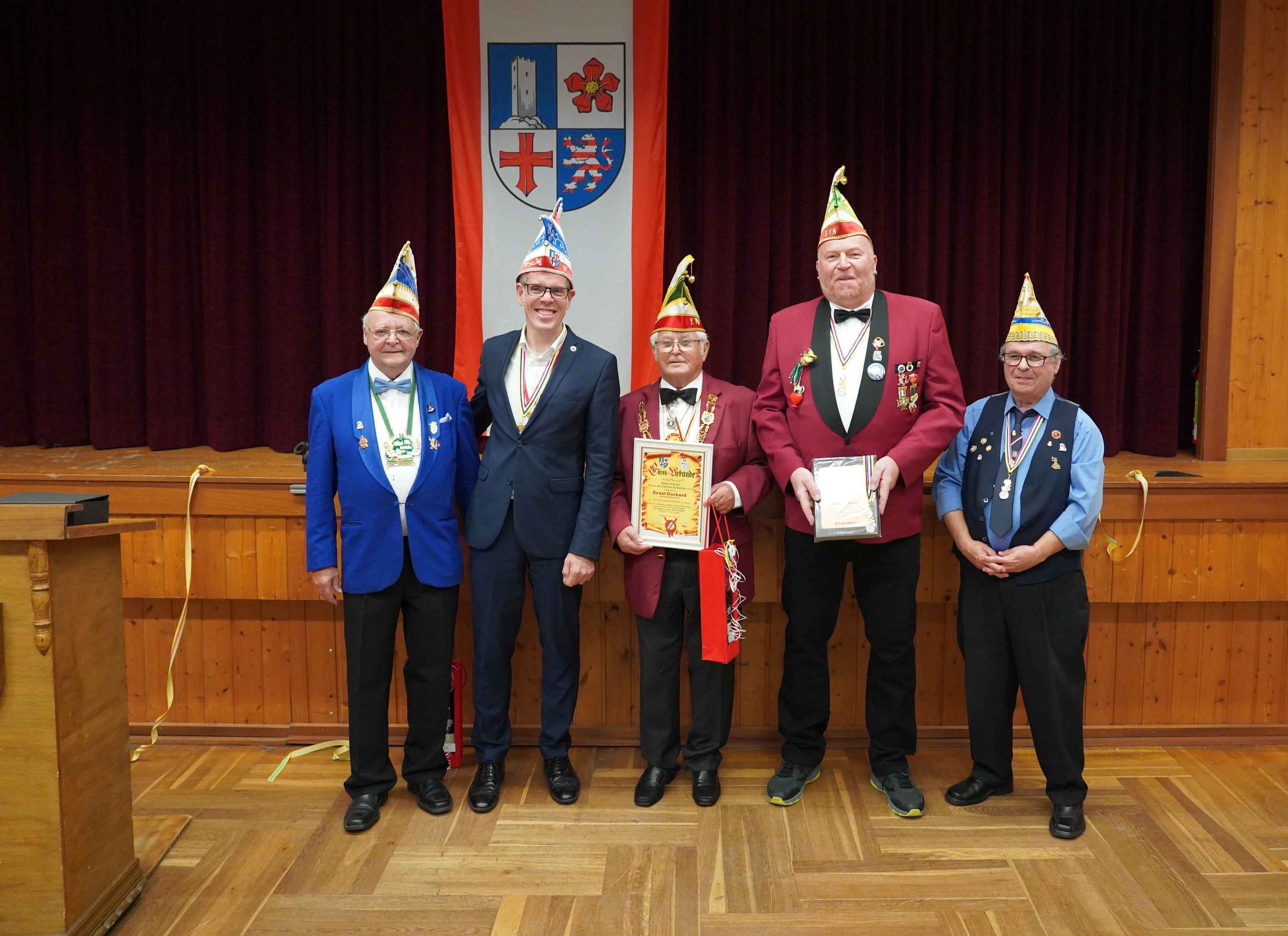 Im Zwingenberger Adlersaal steht Landrat Christian Engelhardt zusammen mit den von ihm geehrten Ernst Gerhard und MArkus Kropp sowie Ulrich Knorr und Siegmund Mendyk von NibA vor der Bühne. Alle tragen wie an Fastnacht üblich Narrenkappen.