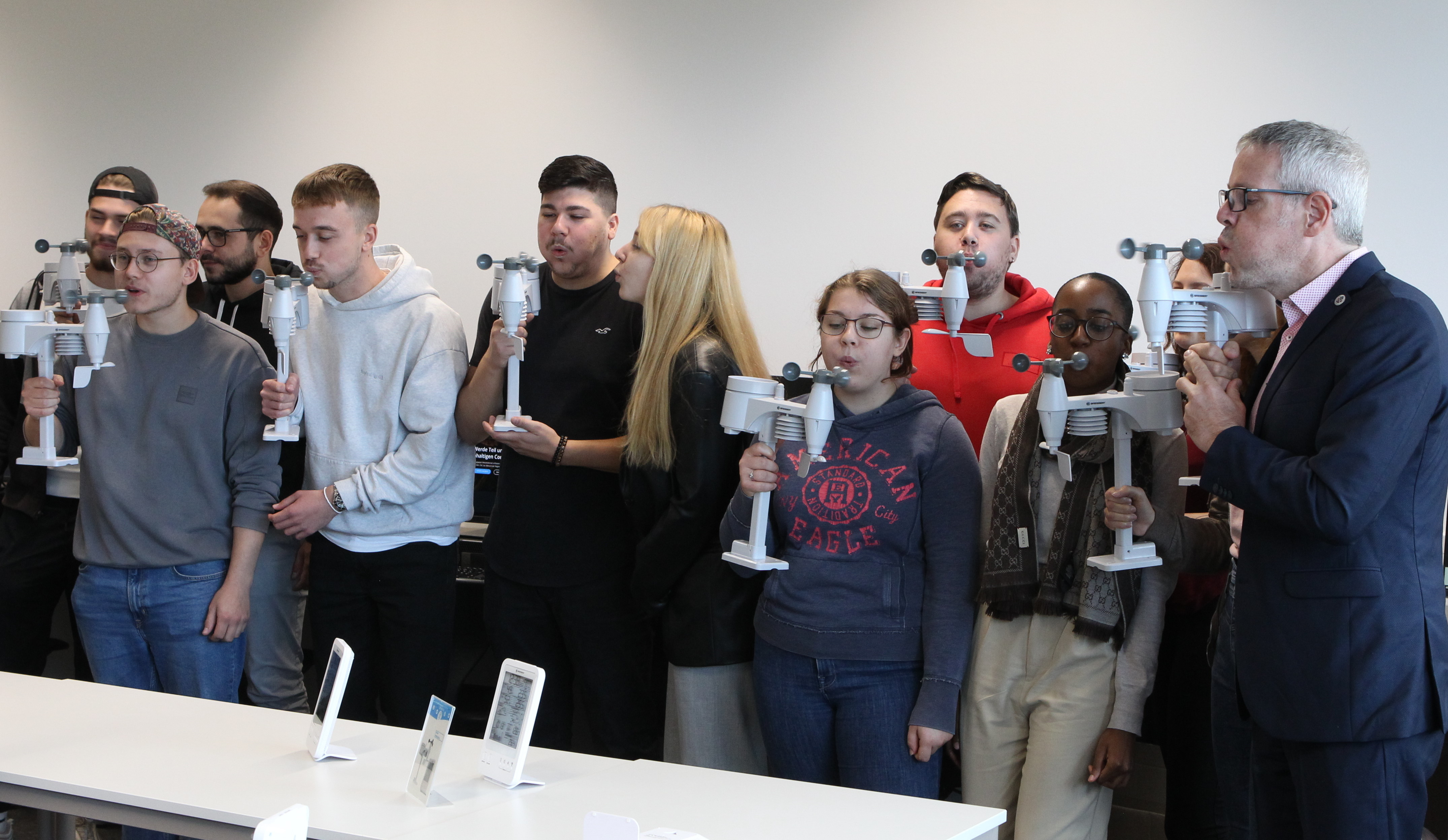 Landrat Christian Engelhardt steht mit Schülerinnen und Schüler der Karl-Kübel-Schule Bensheim in einem Klassenzimmer. Sie testen die Funktion der Windsensoren, indem sie sie in Händen halten und dagegen pusten.