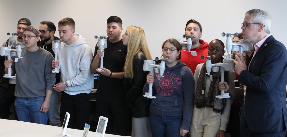 Landrat Christian Engelhardt steht mit Schülerinnen und Schüler der Karl-Kübel-Schule Bensheim in einem Klassenzimmer. Sie testen die Funktion der Windsensoren, indem sie sie in Händen halten und dagegen pusten.