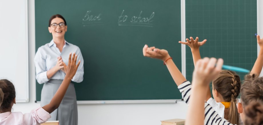 back view of multicultural pupils with hands in air, and teacher standing at chalkboard with back to school inscription, horizontal image