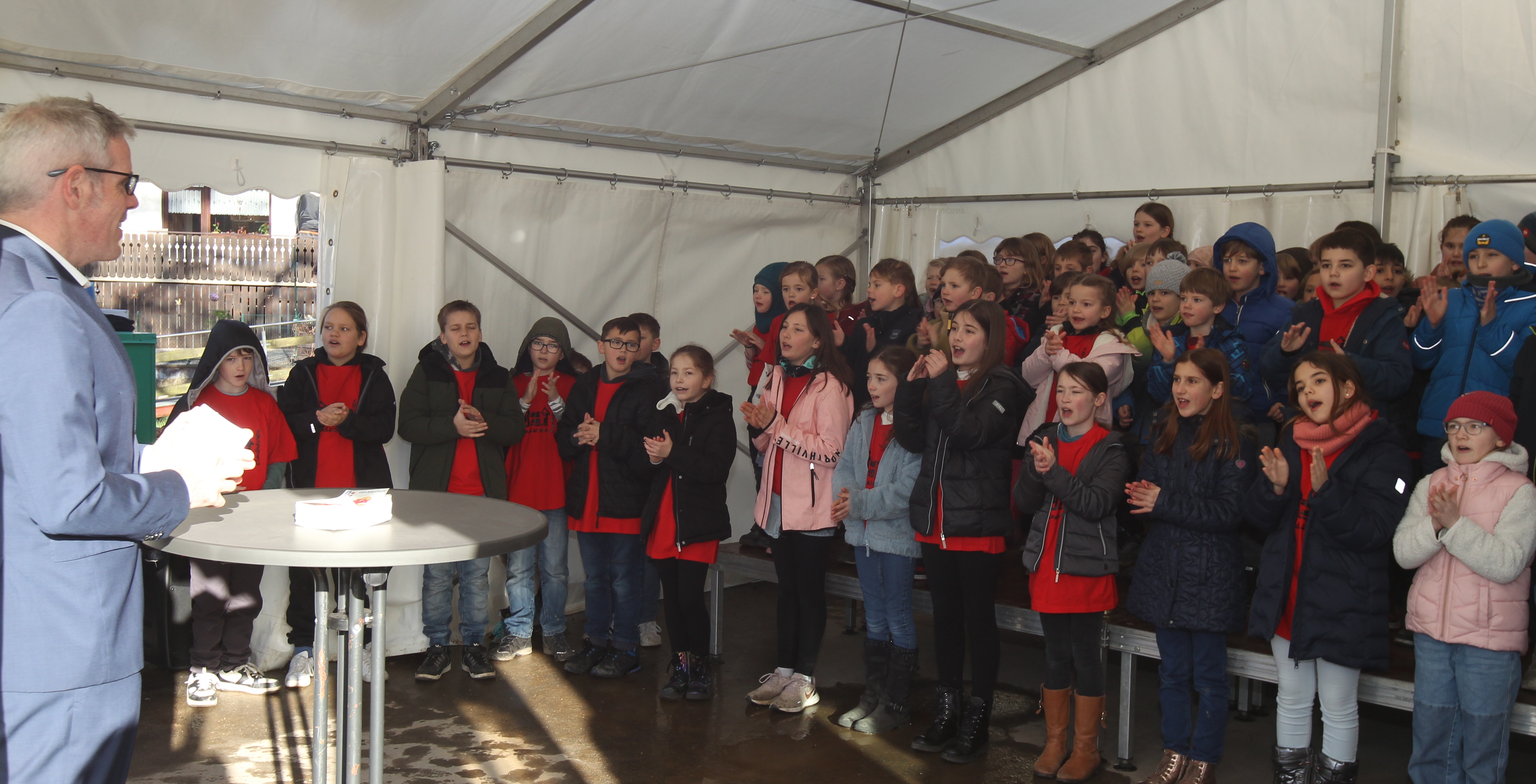 Landrat Christian Engelhardt weiht gemeinsam mit den Schülerinnen und Schülern der Grundschule in Elmshausen die neuen Betreuungsräume ein. Dabei ist er auf dem Bild vor einer Gruppe singender und klatschender Grundschulkinder zu sehen.