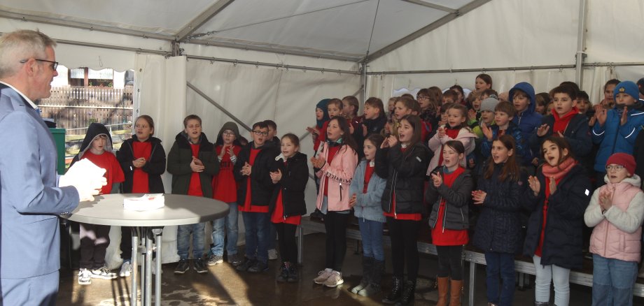 Landrat Christian Engelhardt weiht gemeinsam mit den Schülerinnen und Schülern der Grundschule in Elmshausen die neuen Betreuungsräume ein. Dabei ist er auf dem Bild vor einer Gruppe singender und klatschender Grundschulkinder zu sehen.