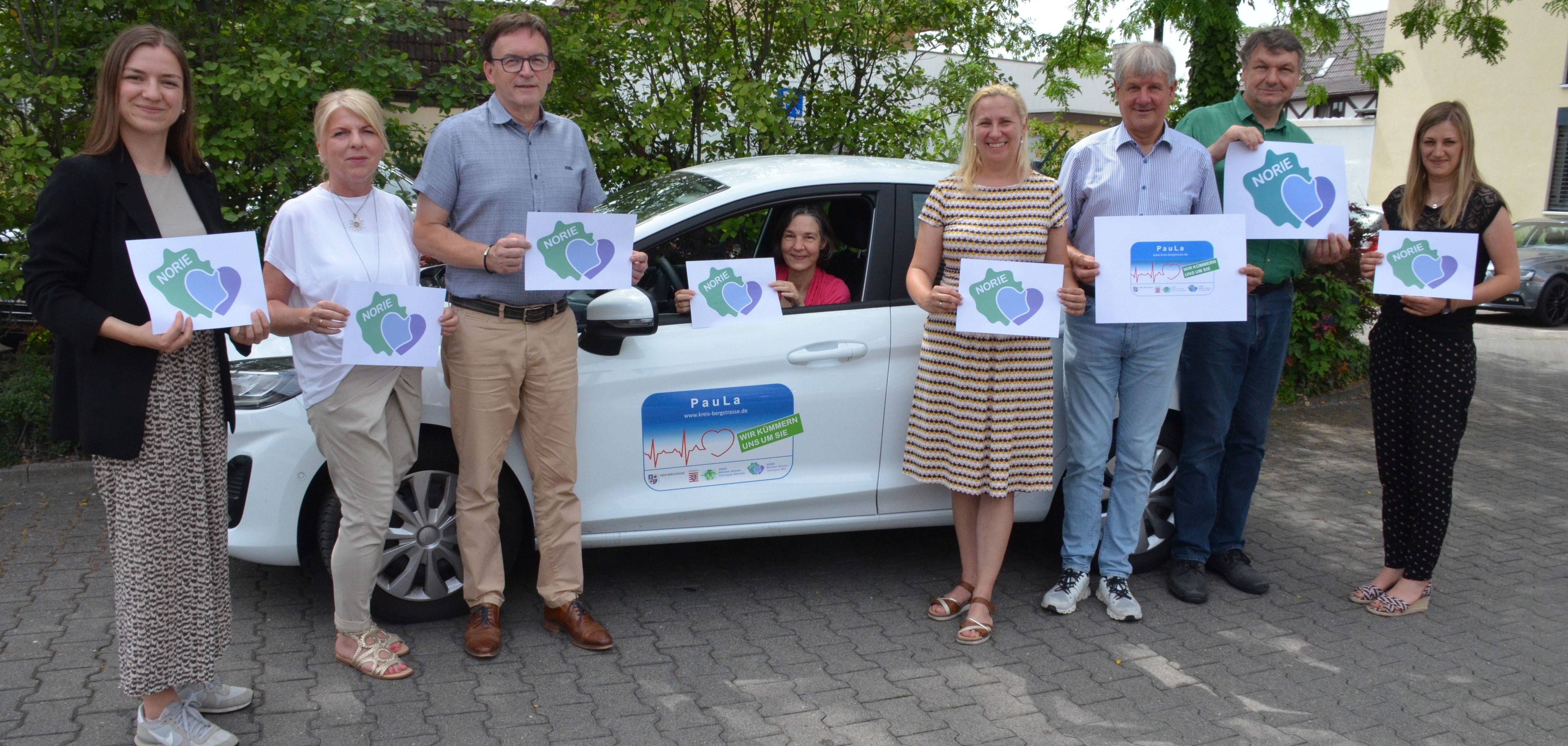 Die Erste Kreisbeigeordnete Diana Stolz (4. v. re.) sowie Vertreterinnen und Vertreter der NORIE-Kommunen präsentieren das neue Logo des Netzwerkes Ortsnahe Versorgung Ried (v.r.n.l.): die Gesundheitskoordinatorin des Kreises Bergstraße Kathleen Wietholtz, der Bürgermeister von Biblis Volker Scheib, der Bürgermeister von Einhausen Helmut Glanzner, die psychosoziale Fachkraft auf dem Land Christina Adler-Schäfer (im Auto sitzend), der Fachbereichsleiter Finanzen und Soziales der Stadt Lorsch Ralf Kleisinger, die Fachdienstleiterin Soziales der Stadt Lorsch Sylvia Weber sowie die Fachbereichsleiterin Gesundheitsversorgung beim Kreis Bergstraße Jennifer Jarke. 