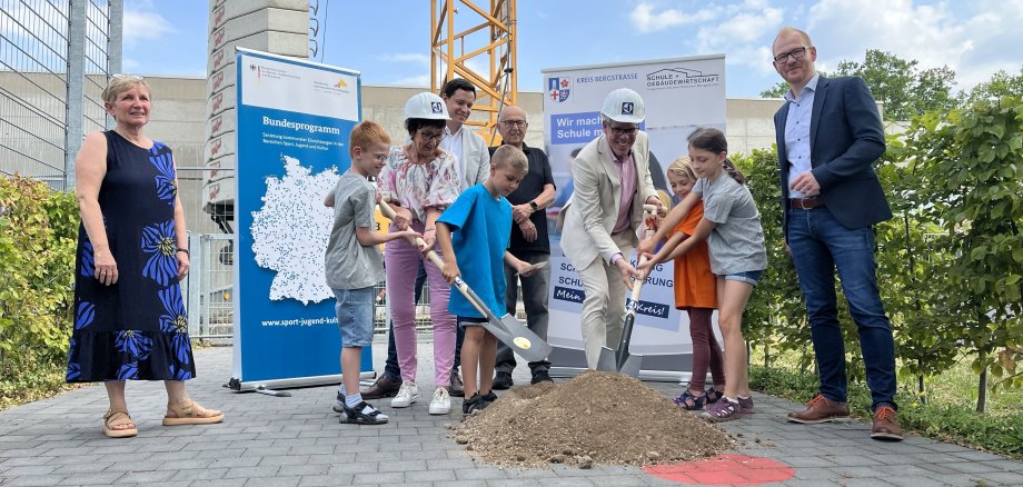 Die Schülerinnen und Schüler des Astrid-Lindgren-Schule unterstützen Landrat Christian Engelhardt (2. v. re.) und Bürgermeisterin Barbara Schader (2. v. li.) beim symbolischen Spatenstich für die neue Sporthalle. Schulleiterin Dagmar Arzberger, der Betriebsleiter des Eigenbetriebs Schule und Gebäudewirtschaft Simon Menden, der Stadtverordnetenvorsteher Franz Siegl und der Betriebsleiter des Eigenbetriebs Schule und Gebäudewirtschaft Johannes Kühn (v.l.n.r.) freuen sich über den Baustart.