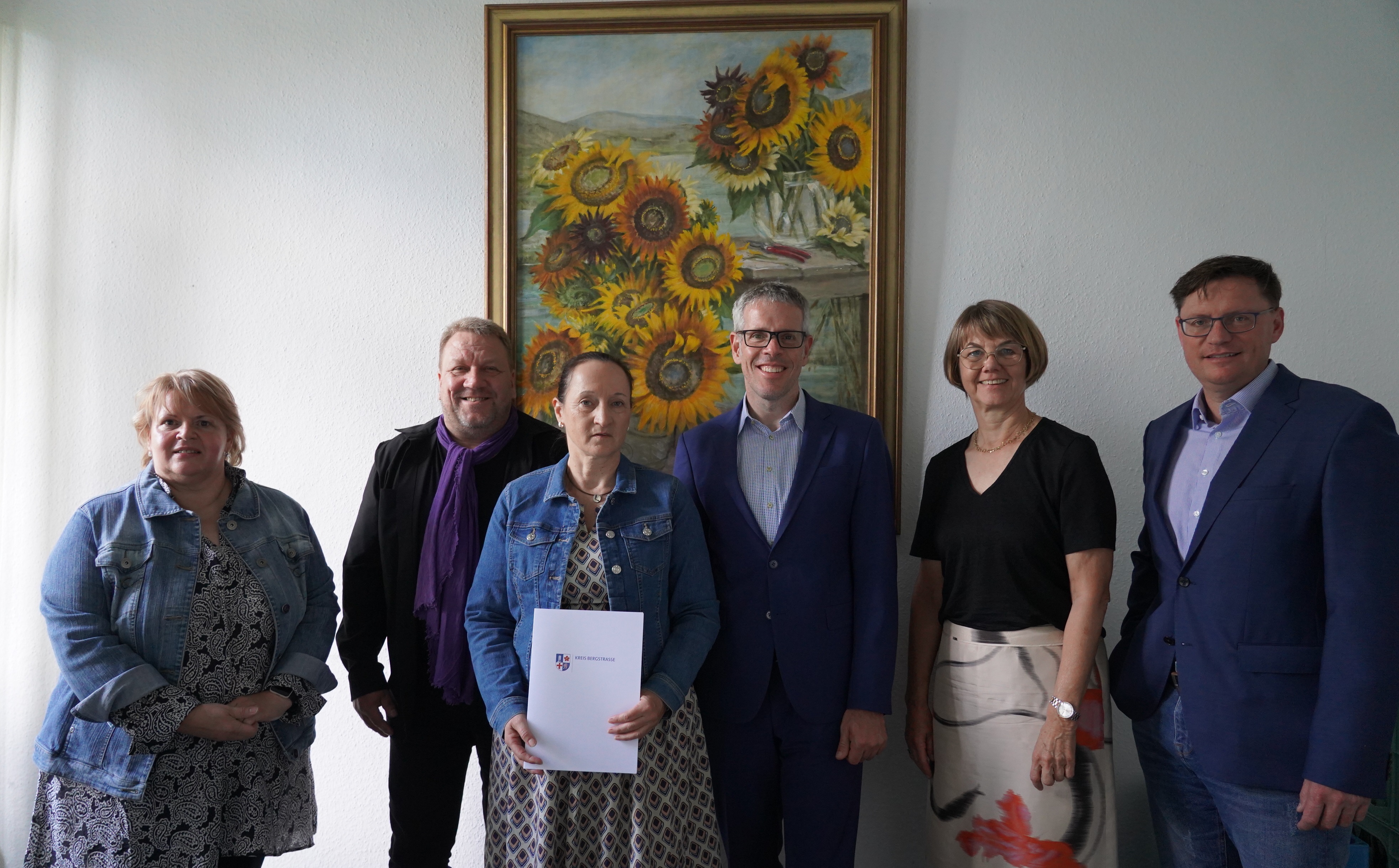 Landrat Christian Engelhardt, die Personalleiterin Barbara Hoffbauer, der Hauptamtliche Kreisbeigeordnete Matthias Schimpf, der Leiter der Abteilung "Soziales" Torsten Bach und Claudia Weinkötz stehen nebeneinander vor einem Sonnenblumengemälde. Frau Weinkötz halt eine Urkunde in den Händen.