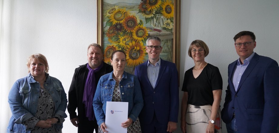 Landrat Christian Engelhardt, die Personalleiterin Barbara Hoffbauer, der Hauptamtliche Kreisbeigeordnete Matthias Schimpf, der Leiter der Abteilung "Soziales" Torsten Bach und Claudia Weinkötz stehen nebeneinander vor einem Sonnenblumengemälde. Frau Weinkötz halt eine Urkunde in den Händen.