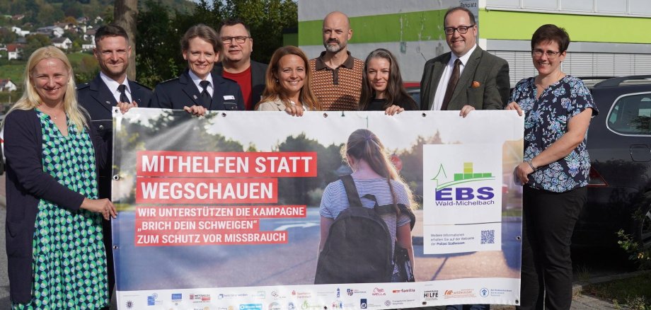 Die Erste Kreisbeigeordnete und Jugenddezernentin Diana Stolz, der Bürgermeister Wald-Michelbachs, Dr. Sascha Weber, die stellvertretende Leiterin der Eugen-Bachmann-Schule, Marina Köhler, der Jugendkoordinator der Polizeidirektion Bergstraße, Johannes Emanuel Hofmann, die Leiterin der Polizeidirektion Bergstraße, Juliane Ries, der Leiter des Bergsträßer Jugendamtes, Kai Kuhnert, die Präventionslehrerin der EBS, Lisa Perez, der Schulpsychologe Tobias Knapp und die Kriminaloberkommissarin Lucia Mitsch stehen mit dem Banner der Kampagne „Brich Dein Schwiegen – hinter jedem Missbrauch steckt ein Gesicht“ vor der Eugen-Bachmann-Schule.