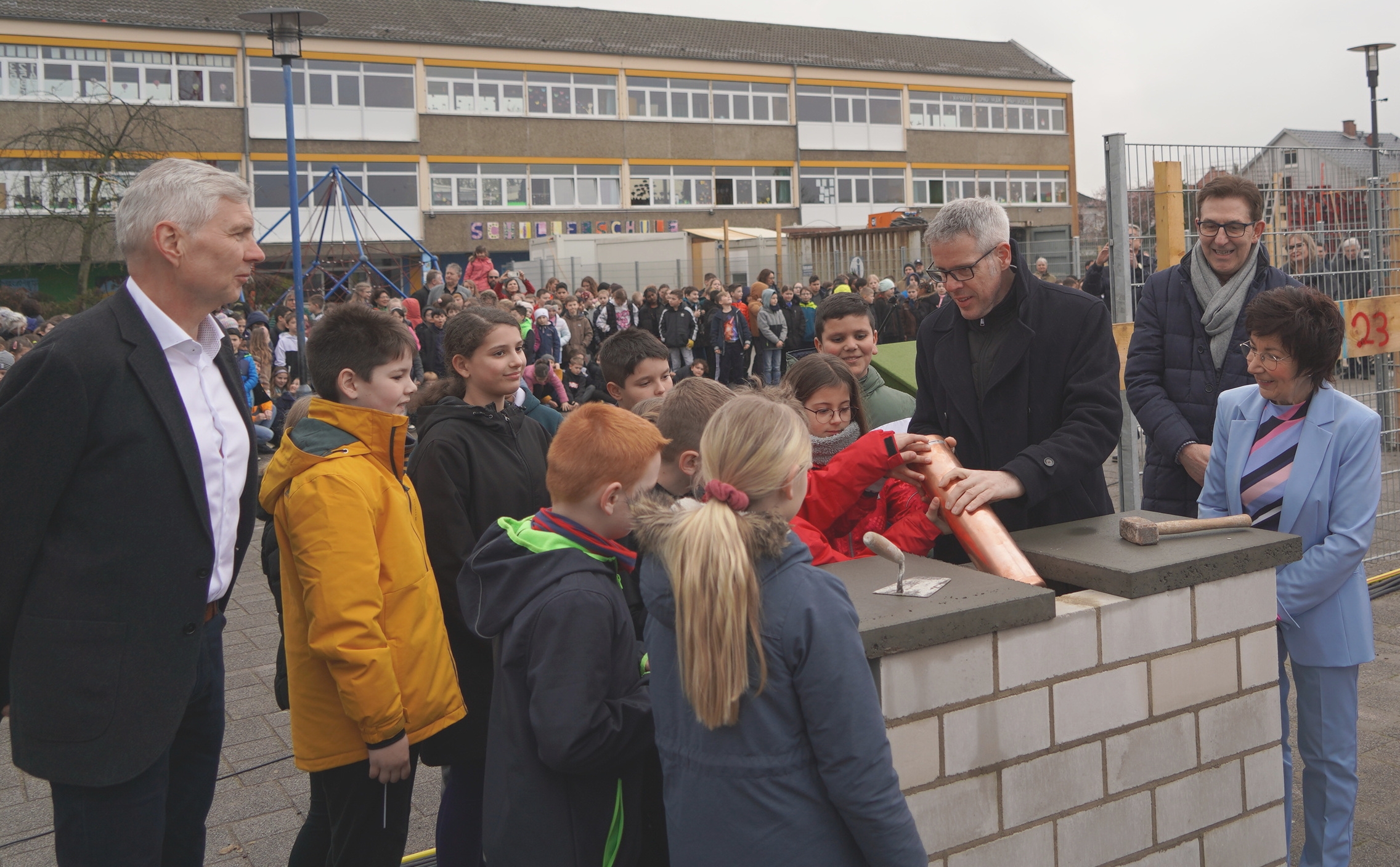 Landrat Christian Engelhardt lässt auf dem Schulhof der Schillerschule Bürstadt gemeinsam mit dem Schulleiter Torsten Wiechmann, der Bürgermeisterin Barbara Schader, dem Architekten Klaus Klinger und den Schulkindern eine Zeitkapsel in die Mauer des Neubaus.