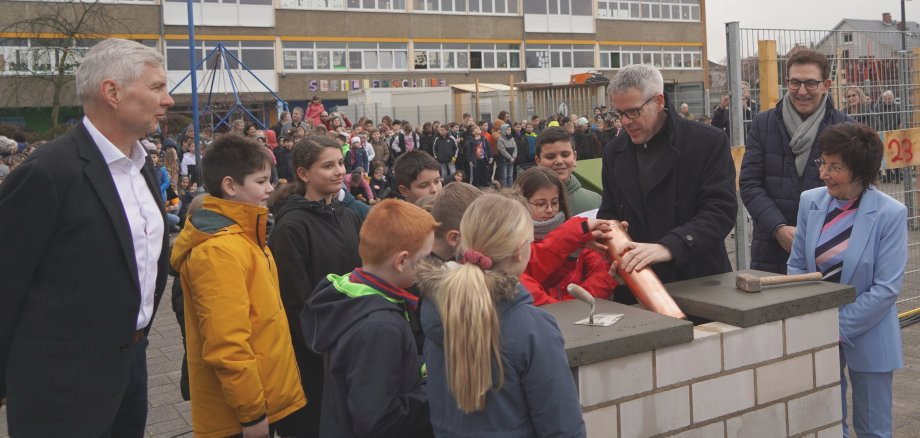 Landrat Christian Engelhardt lässt auf dem Schulhof der Schillerschule Bürstadt gemeinsam mit dem Schulleiter Torsten Wiechmann, der Bürgermeisterin Barbara Schader, dem Architekten Klaus Klinger und den Schulkindern eine Zeitkapsel in die Mauer des Neubaus.