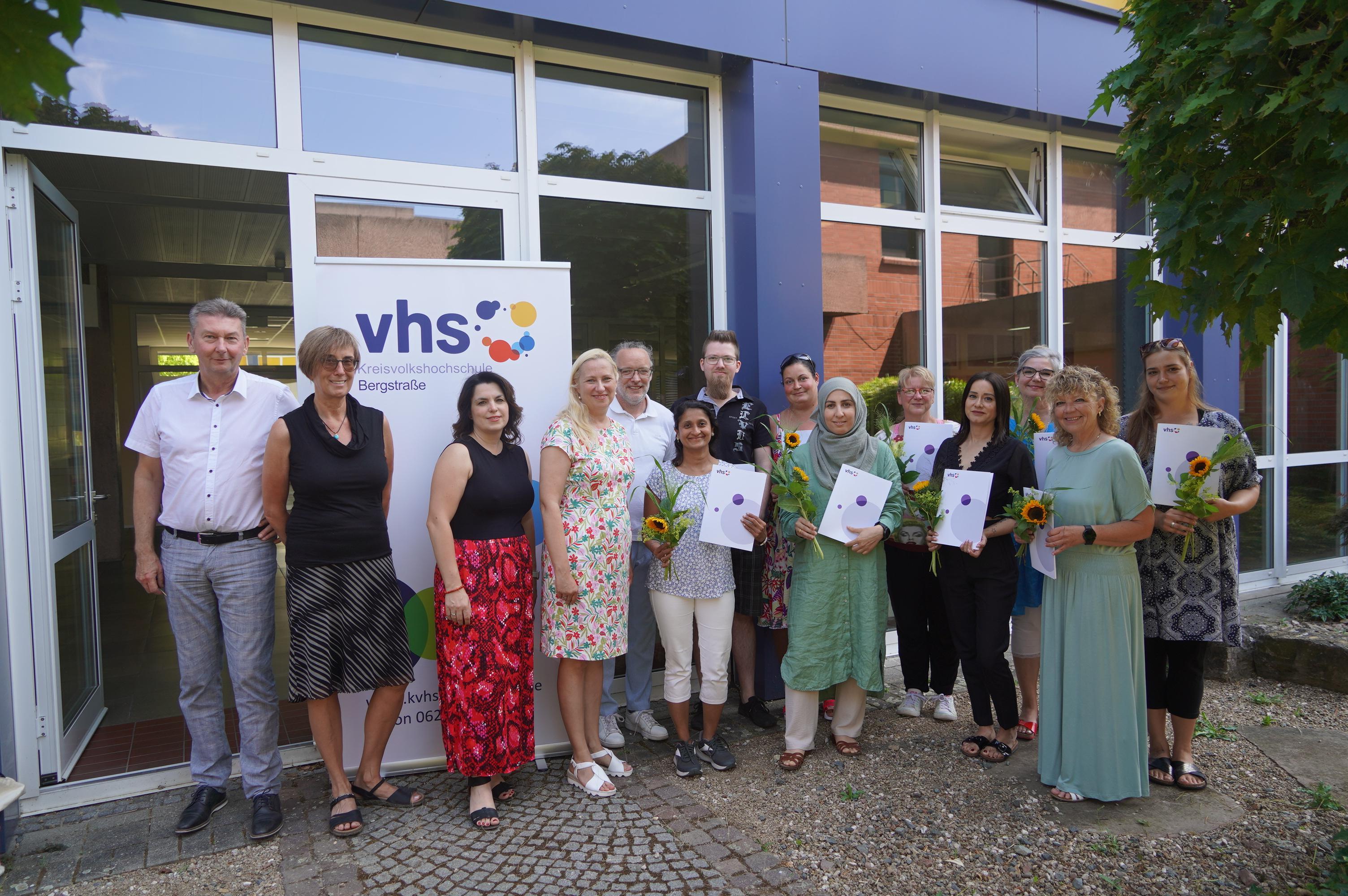 Die Absolventinnen und Absolventen des 5. Landeslehrgangs für Betreuungskräfte aus dem Pakt für den Ganztag Die Erste Kreisbeigeordnete Diana Stolz steht gemeinsam mit dem kommissarischen Schulleiter der ESS Martin Gonnermann, Claudia Blume vom Eigenbetrieb Schule und Gebäudewirtschaft Kreis Bergstraße, der Fachbereichsleiterin der Kreisvolkshochschule Bergstraße Melanie Knauf, dem Kursleiter Christian Hörnle und den Absolventinnen und Absolventen des fünften Landeslehrgangs für Betreuungskräfte aus dem Pakt für den Ganztag vor der ESS. Die Teilnehmenden halten jeweils ihr Zertifikat und eine Sonnenblume in den Händen.