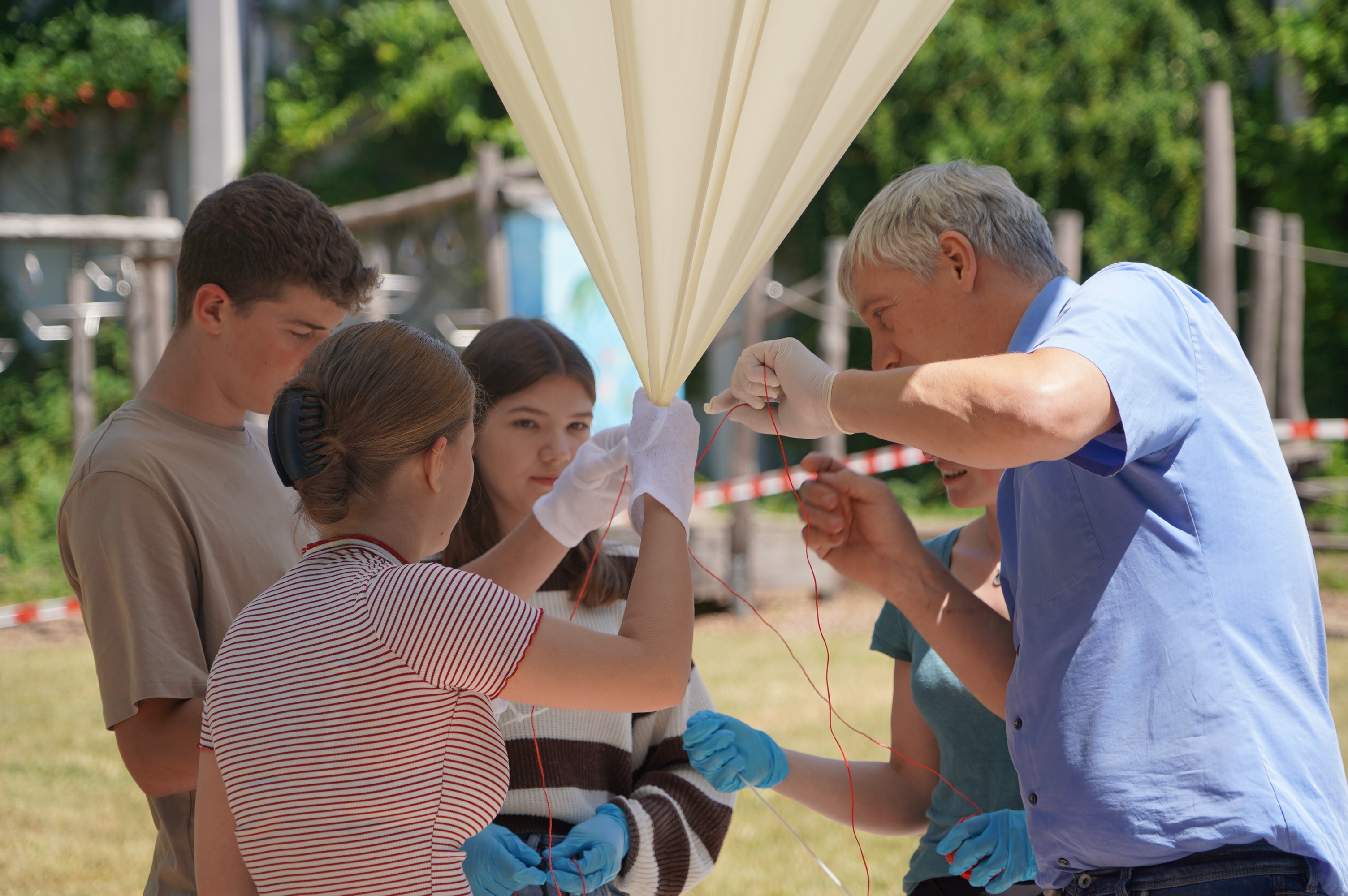 Die Schülerinnen und Schüler des Naturwissenschaftskurses, Kurslehrerin Eva Nagel und der Referent für digitale Bildung und Leiter des digi_space des Kreises Bergstraße Jan Fuchs befestigen die Sonde am Ballon. Die Schülerinnen und Schüler des Naturwissenschaftskurses, Kurslehrerin Eva Nagel und der Referent für digitale Bildung und Leiter des digi_space des Kreises Bergstraße Jan Fuchs halten gemeinsam den mit Helium gefüllten, weißen Ballon und befestigen mit einer roten Kordel die Sonde.