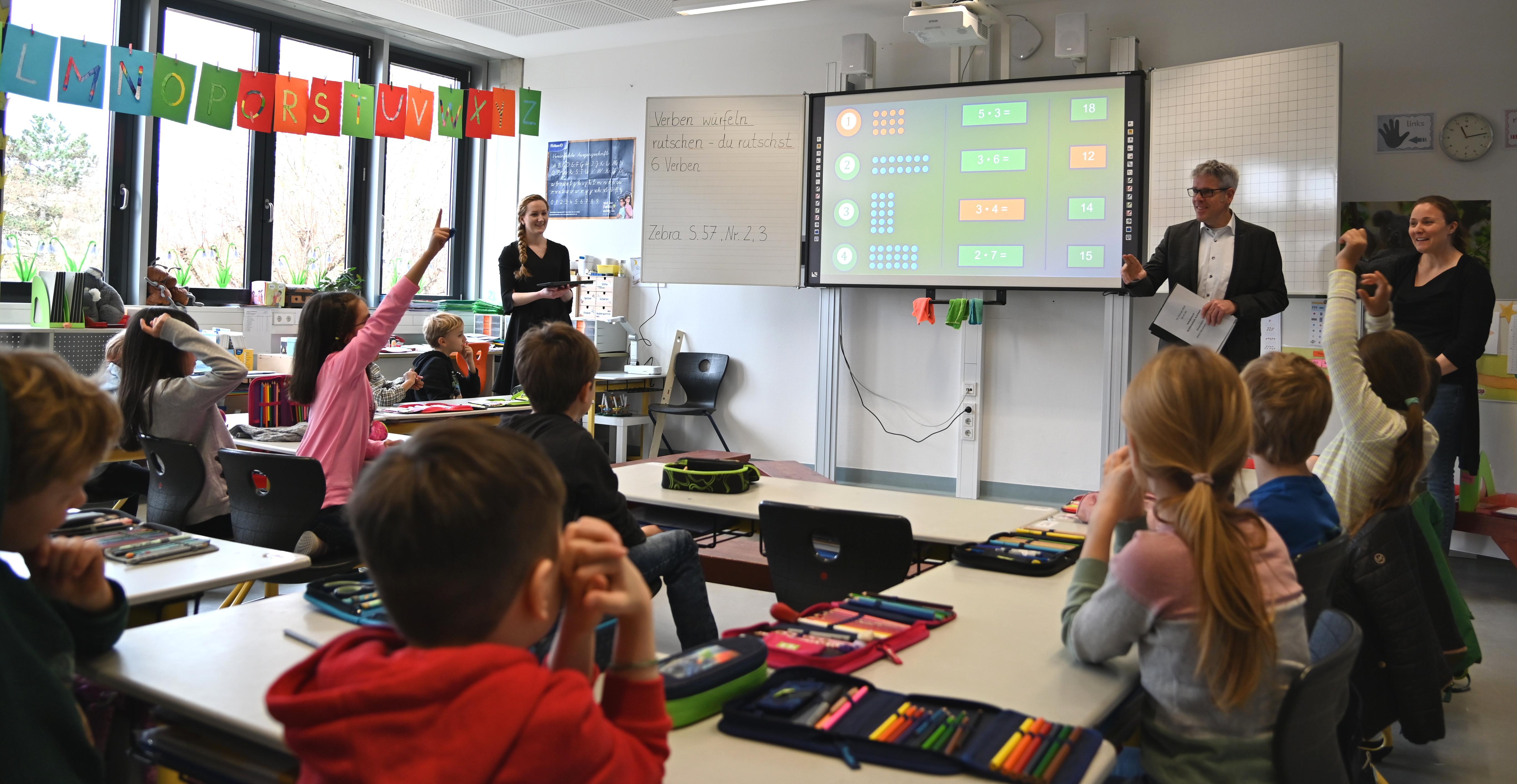 Das Bild zeigt Landrat Christian Engelhardt in einem Klassenzimmer der Schillerschule in Bensheim-Auerbach. Er steht an der Tafel und ist mit Schülerinnen und Schülern im Gespräch, von denen sich einige melden.