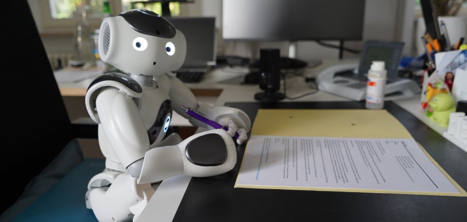 Der kleine Roboter "Nao" sitzt am Schreibtisch des Landrats und unterzeichnet ein Dokument.