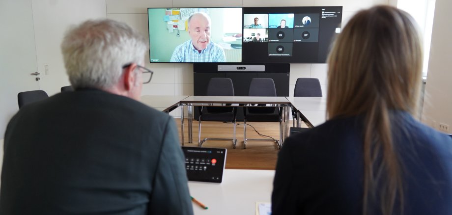 Landrat Christian Engelhardt sitzt gemeinsam mit der Gesundheitskoordinatorin Kathleen Wietholtz mit dem Rücken zur Kamera in der hybriden Pressekonferenz vor einem Monitor.