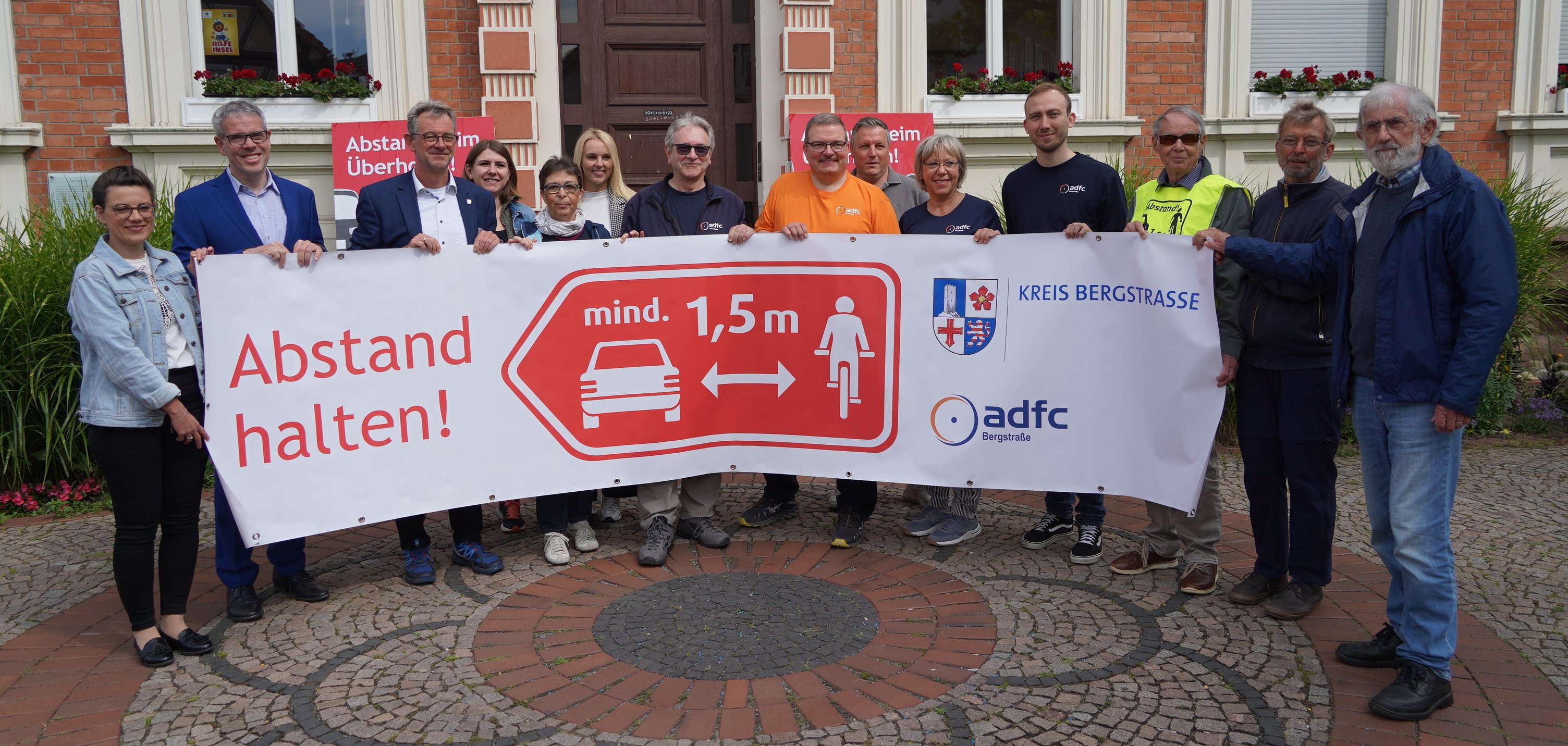 Landrat Christian Engelhardt steht gemeinsam mit dem Bürgermeister der Stadt Lorsch, Christian Schönung, der in der Kreisverwaltung für den Radverkehr zuständigen Sachbearbeiterin Lisa-Marie Riemann, der ersten Vorsitzenden des ADFC, Anette Seip, dem zweiten Vorsitzenden des ADFC, Friedhelm Höcker sowie weiteren Vertreterinnen und Vertretern der Kooperationspartner sowie teilnehmenden Kommunen beim Aktionsauftakt in Lorsch vor dem Rathaus und hält ein AKtionsbanner hoch.