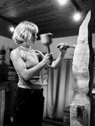 Eine Bildhauerin steht mit Nagel und Meißel in den Händen an einem großen Stein und meißelt.