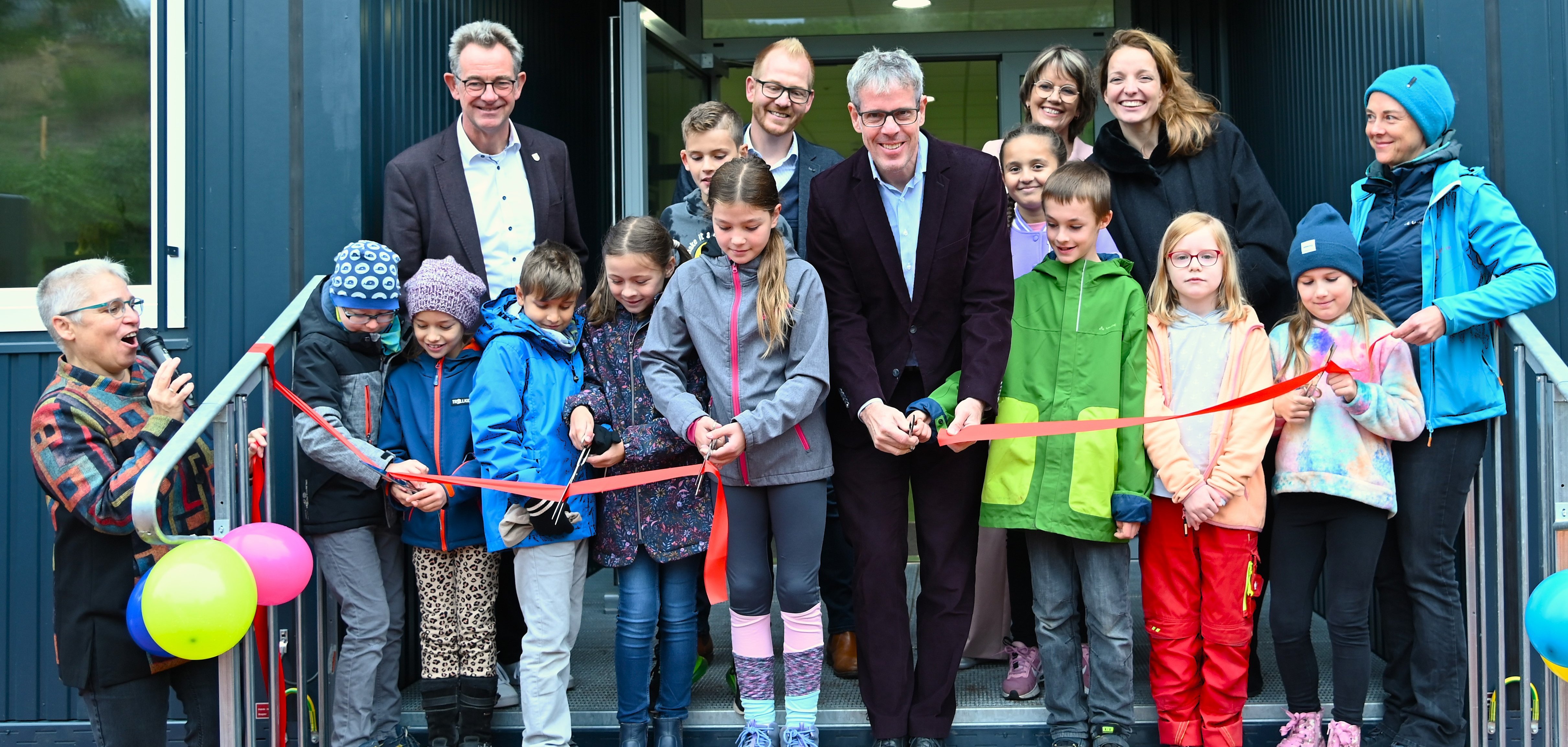Landrat Christian Engelhardt schneidet gemeinsam mit Schulleiterin Jutta Rothfritz, Vertreterinnen und Vertretern aus Politik, Bildung und des Eigenbetriebs Schule und Gebäudewirtschaft sowie der Schulgemeinde ein rotes Band vor dem neuen Schulgebäude durch.