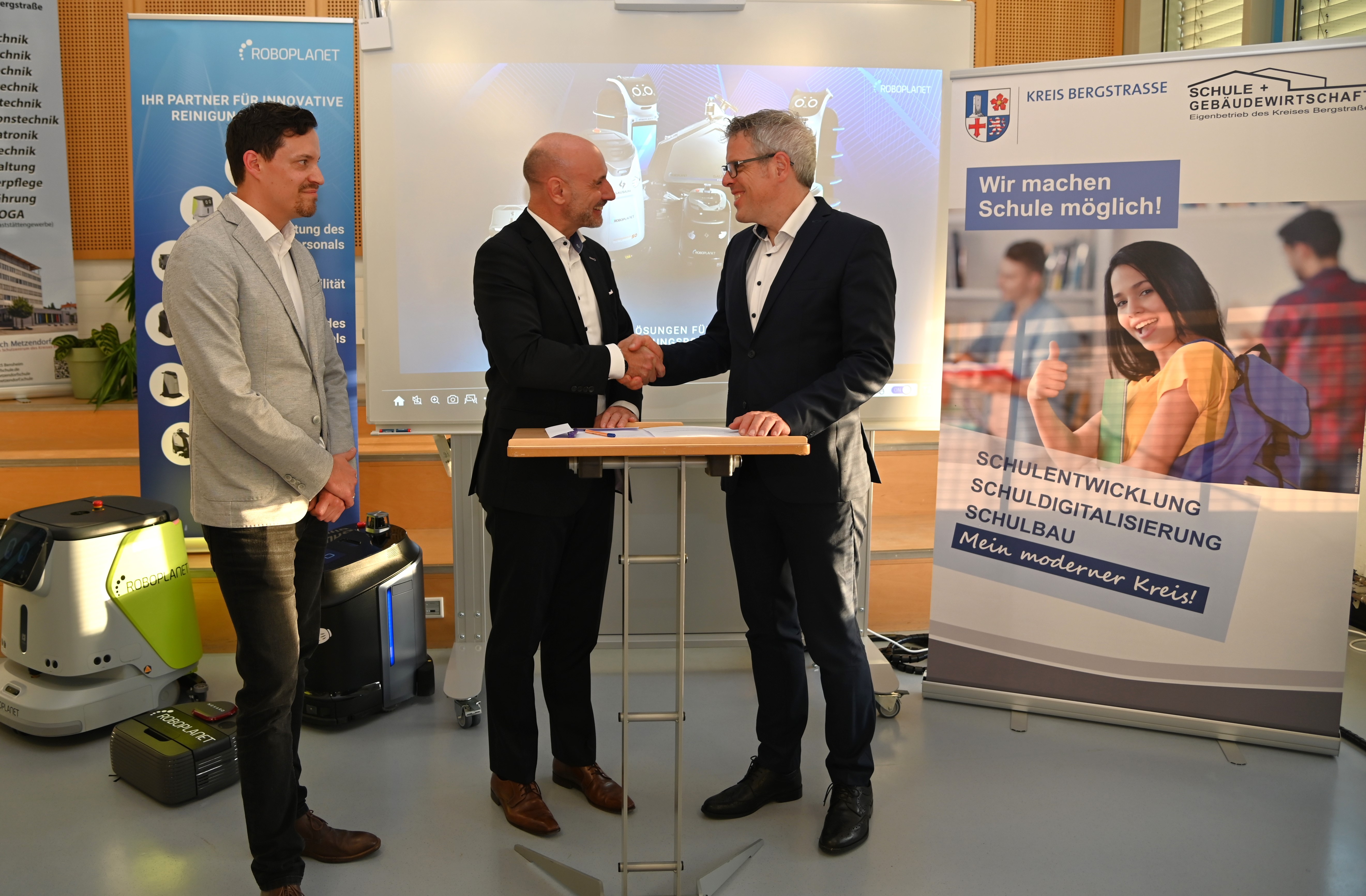 Landrat Christian Engelhardt und André Pflüger, Geschäftsführer der Wackler Service Group GmbH, stehen vor Aufstellern an einem Tisch und schütteln die Hand. Links daneben steht der kaufmännische Betriebsleiter des Eigenbetriebs Schule und Gebäudewirtschaft, Simon Menden, neben einem der Putzroboter.