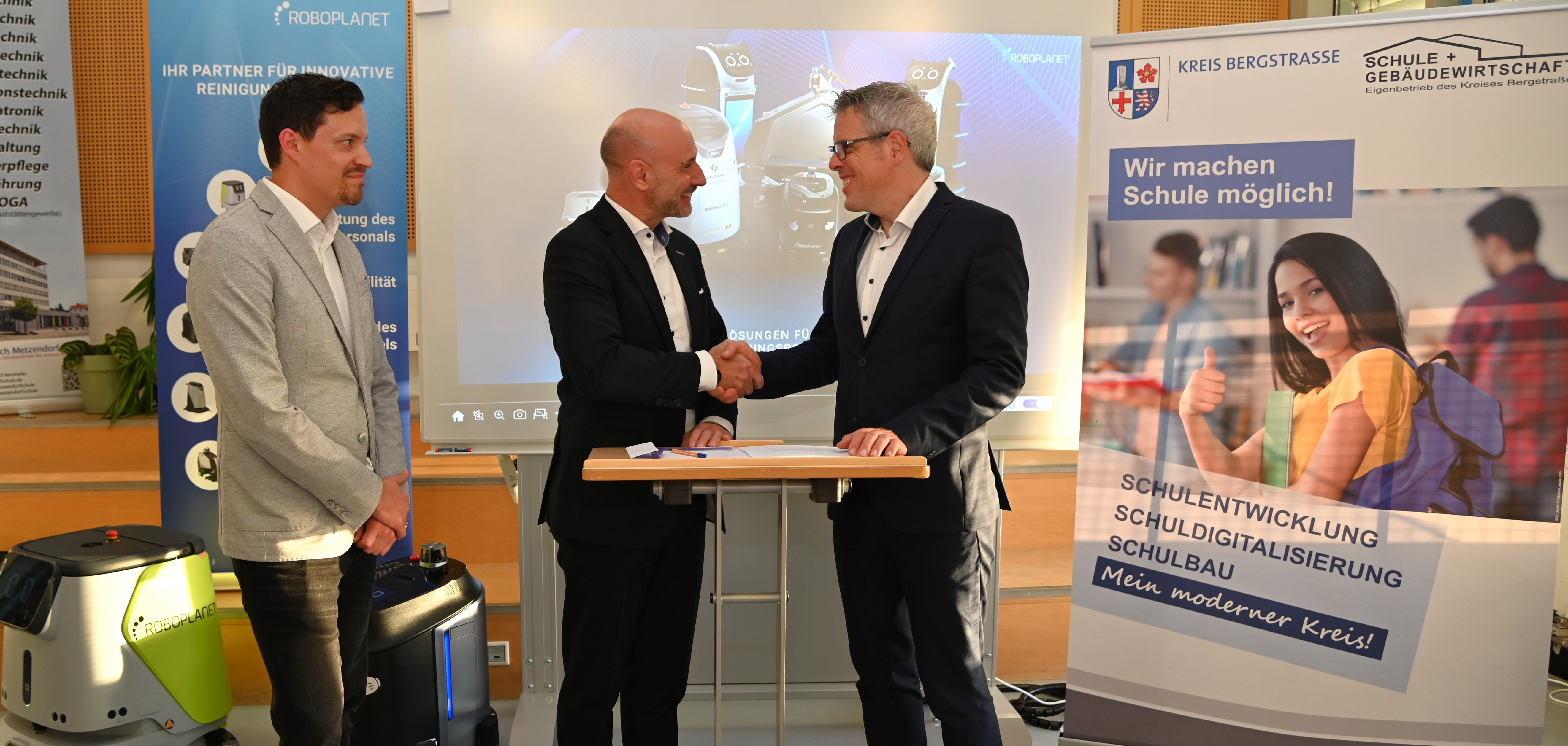 Landrat Christian Engelhardt und André Pflüger, Geschäftsführer der Wackler Service Group GmbH, stehen vor Aufstellern an einem Tisch und schütteln die Hand. Links daneben steht der kaufmännische Betriebsleiter des Eigenbetriebs Schule und Gebäudewirtschaft, Simon Menden, neben einem der Putzroboter.
