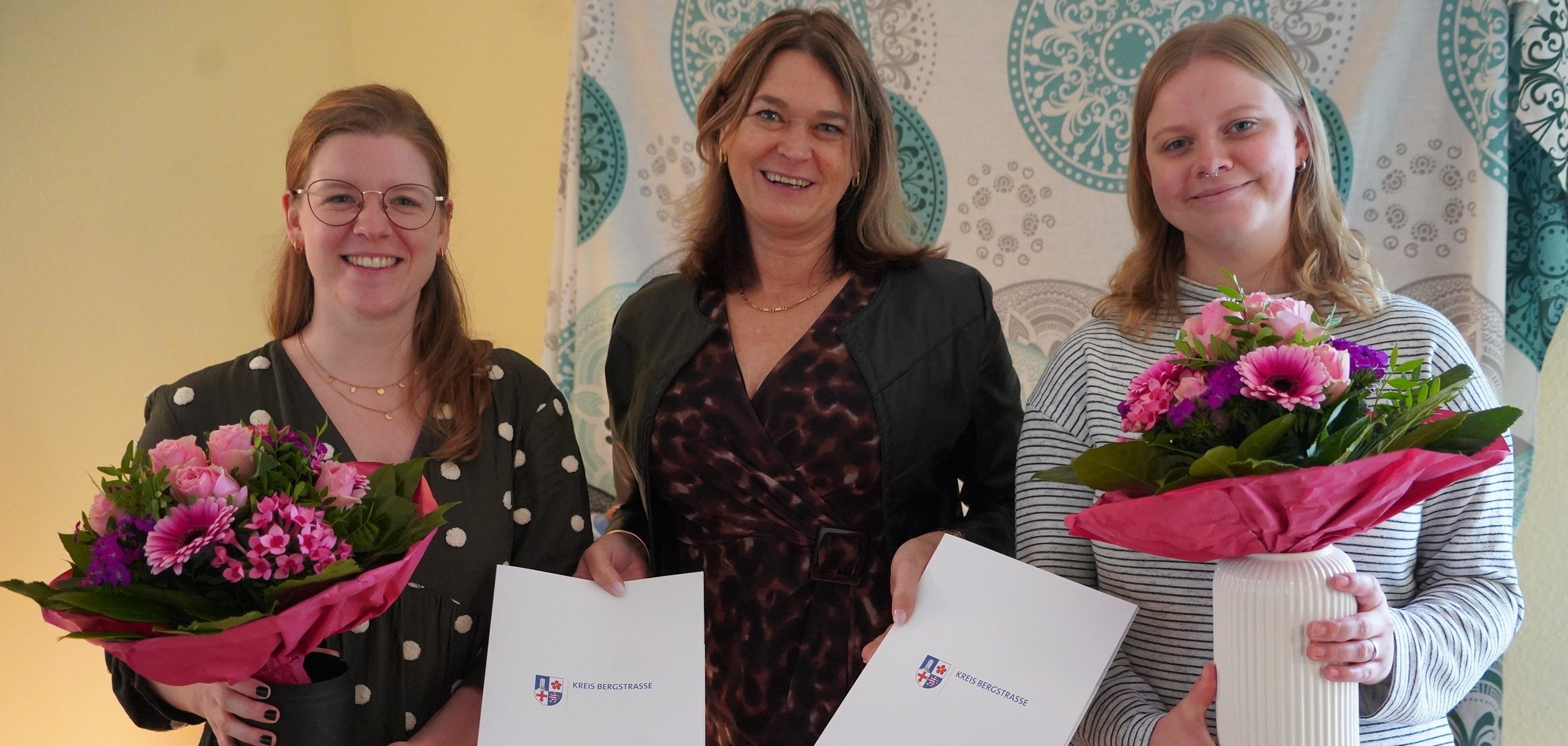 Die Erste Kreisbeigeordnete und Gesundheitsdezernentin Angelika Beckenbach steht mit den Hebammen Annika Brock (links) und Hanna Kriegs im Geburtshaus. Beide halten jeweils einen rosa Blumenstrauß in den Händen. Die Gesundheitsdezernentin hält die Förderbescheide über die Erstausstattung im Nachsorgebereich in den Händen.