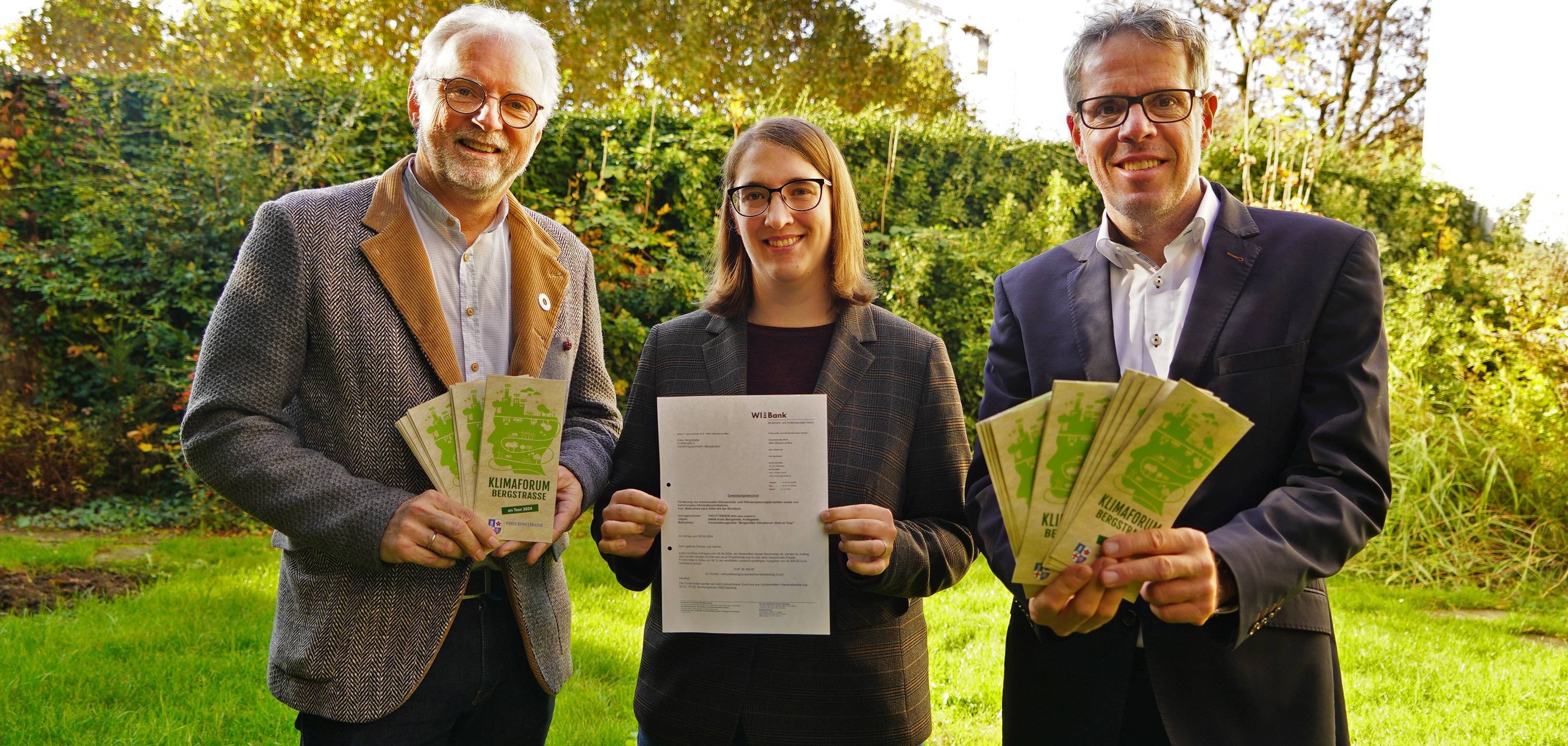 Landrat Christian Engelhardt, Katrin Heuer und Reiner Pfuhl vom Klimaschutzteam des Kreises Bergstraße stehen vor einer grünen Hecke und halte den Förderbescheid des HMLU sowie Flyer des Klimaforums on Tour in den Händen.