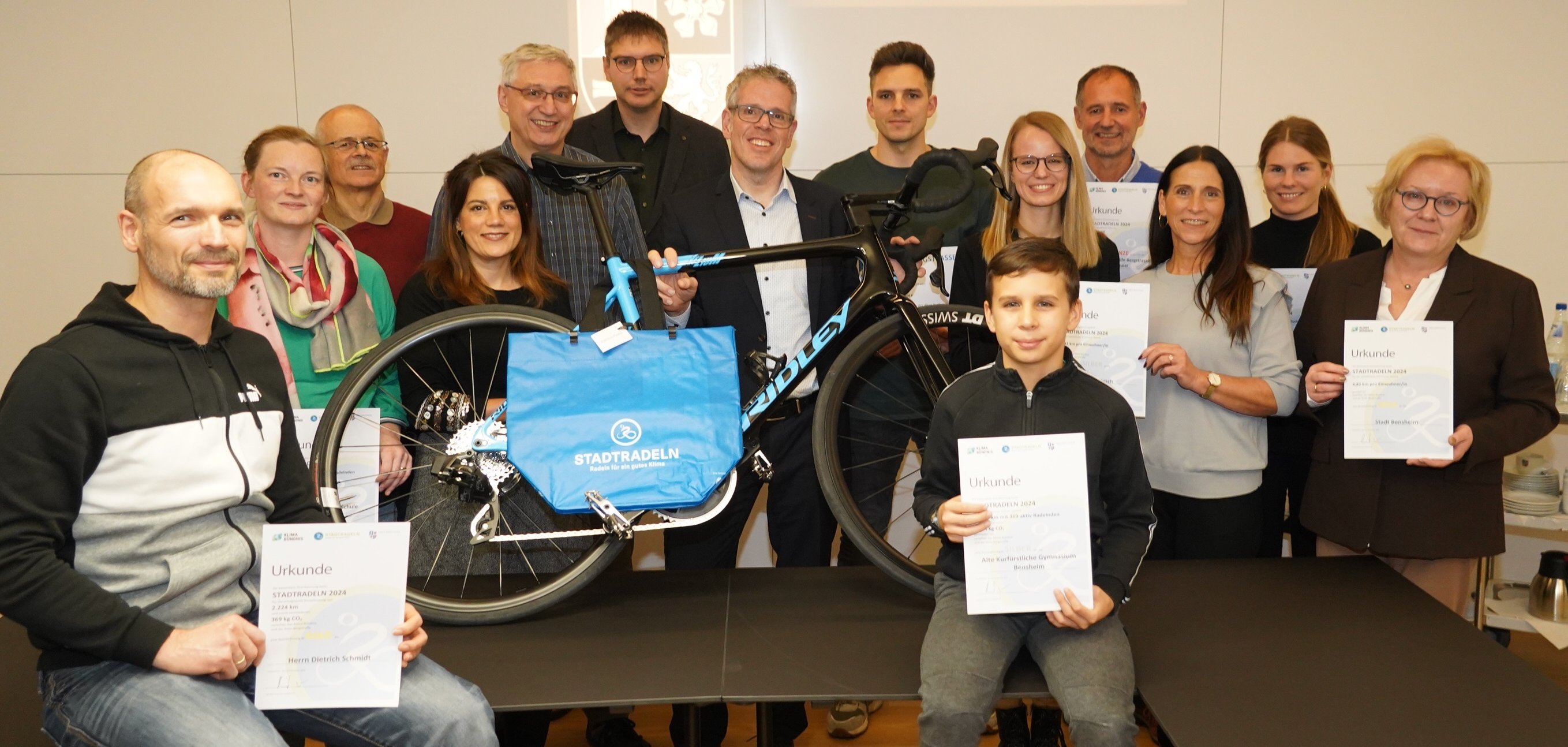 Landrat Christian steht mit den Geehrten sowie Dominik Perleth und Lisa-Marie Riemann von der Abteilung ÖPNV und Mobilität um einen Tisch herum. Auf dem Tisch steht ein Fahrrad. Die Geehrten halten ihre Urkunden in den Händen.