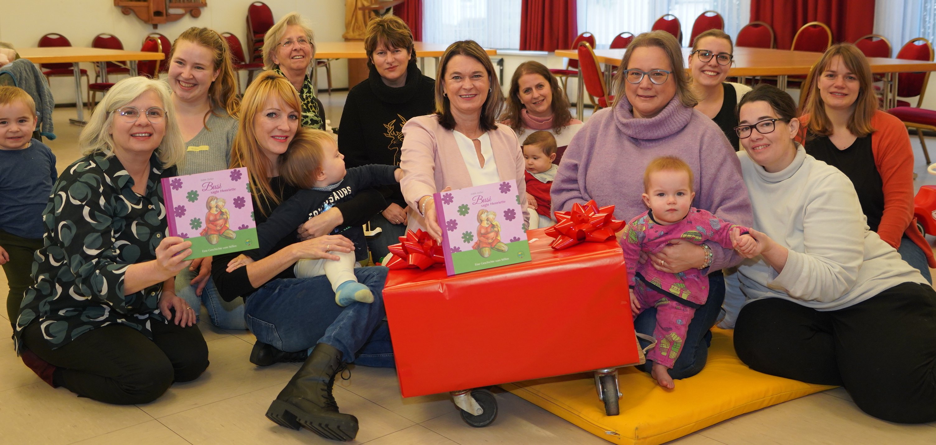 Die Erste Kreisbeigeordnete und Gesundheitsdezernentin Angelika Beckenbach  sitzt gemeinsam mit Georgeta Ensinger und Sieglinde Weimar von den Frühen Hilfen des Kreises Bergstraße, Heike Schön vom Arbeitskreis Jugendzahnpflege, der Projektleiterin Inka Kuusela sowie einigen Besucherinnen mit ihren Kindern im Drop In(klusive)-Treffs im Pfarrzentrum der Katholischen Pfarrgemeinde Sankt Laurentius in Bensheim auf einer Matte auf dem Buden. Vor ihnen stehen die in ein großes Paket eingepackten Stillbücher.